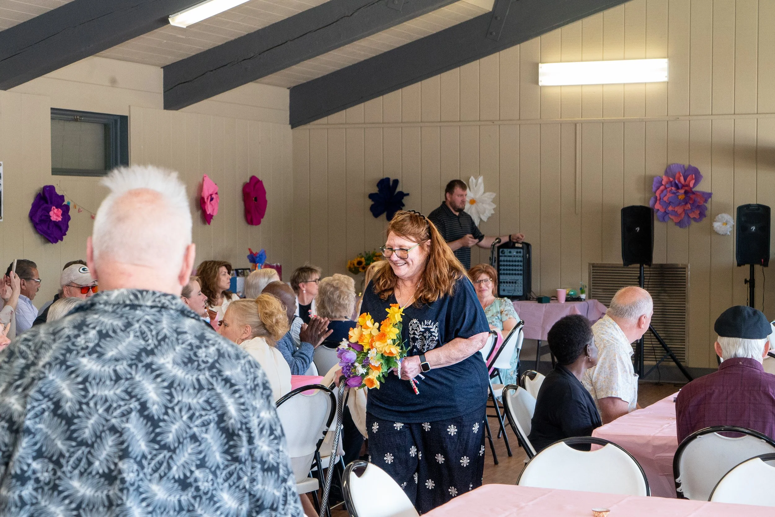 A woman holding a colorful bouquet of flowers, smiling, at a crowded indoor event with decorated walls and a man in the background operating sound equipment.