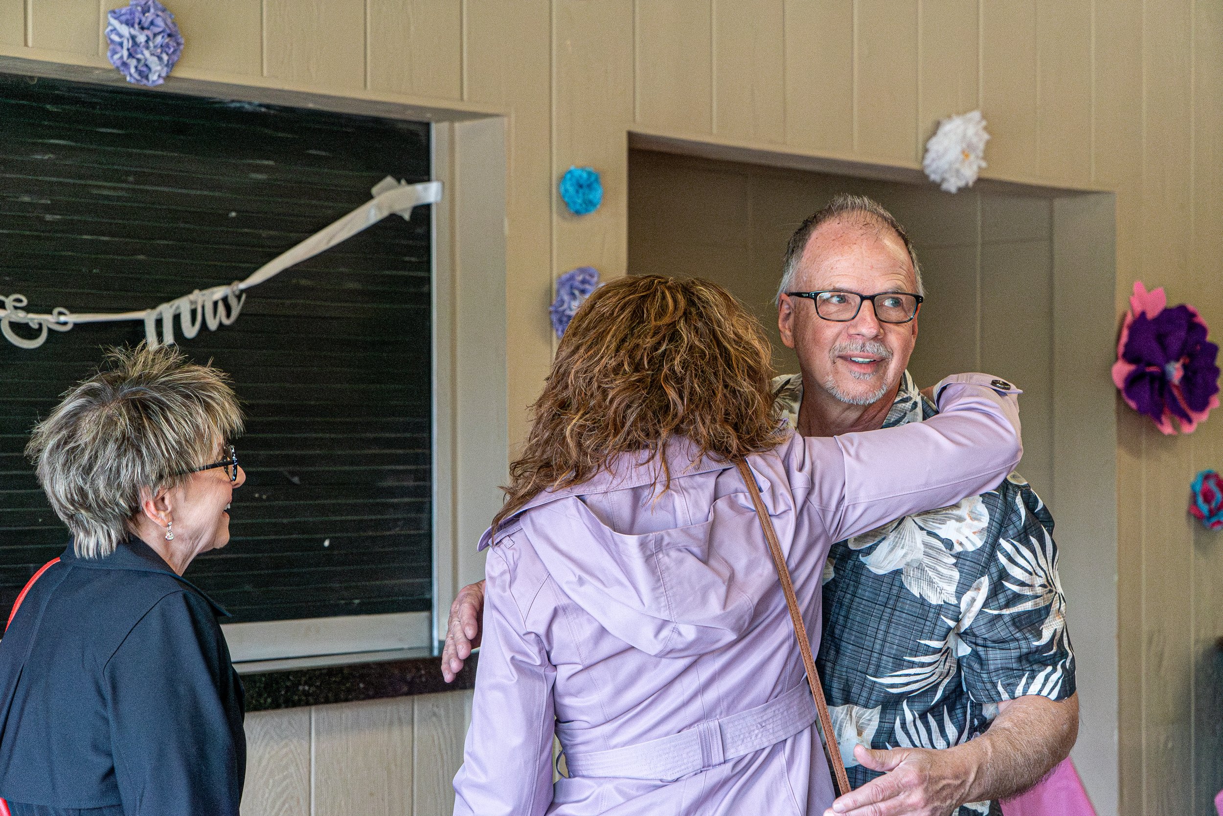 People embracing at a wedding with decorations, including paper flowers and a banner, in an indoor setting.