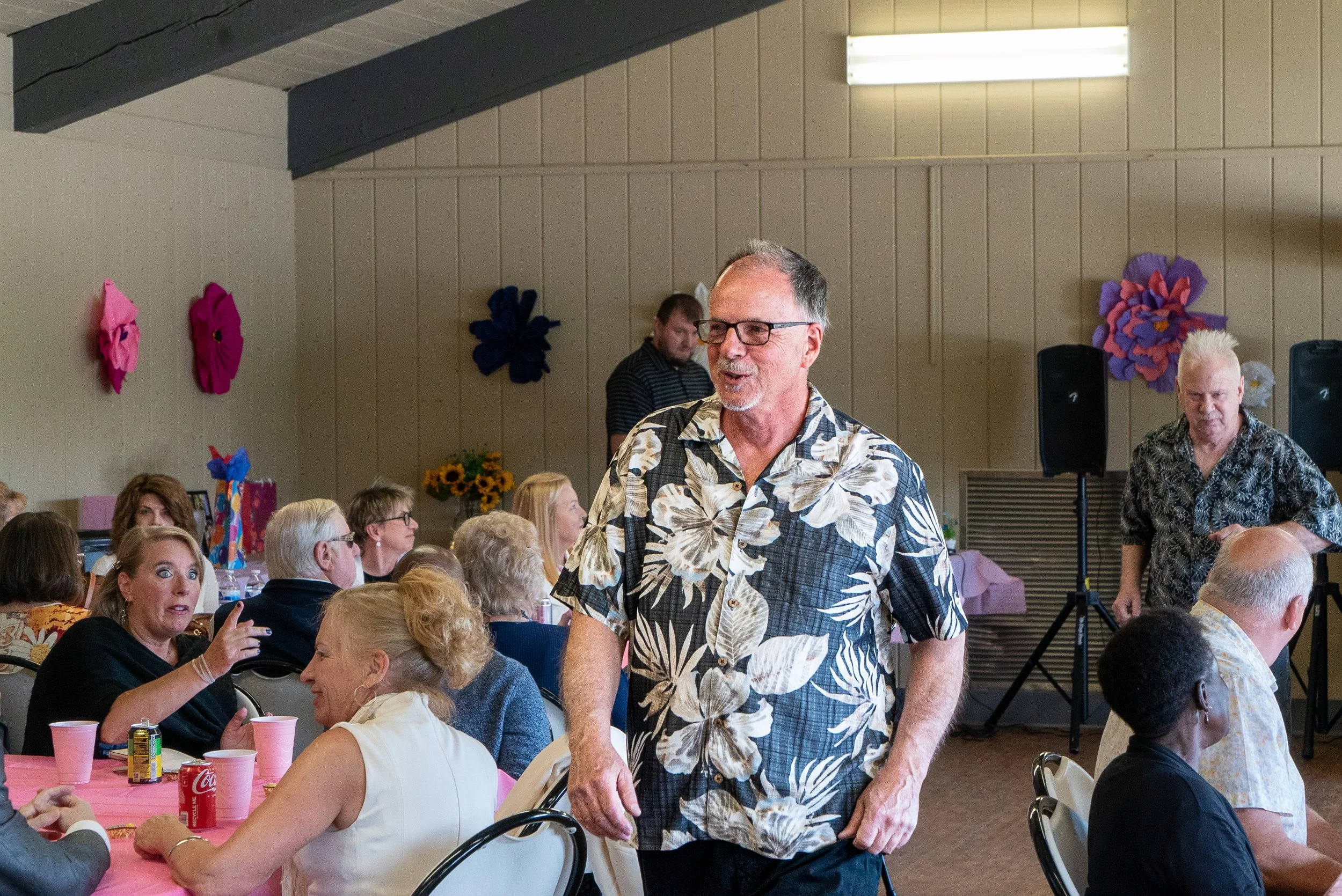 People gathered at a party or celebration, with some sitting at tables and others standing and talking. The room has decorated walls with large colorful paper flowers, and there are tables with pink tablecloths, cups, and food or drinks. The atmosphe