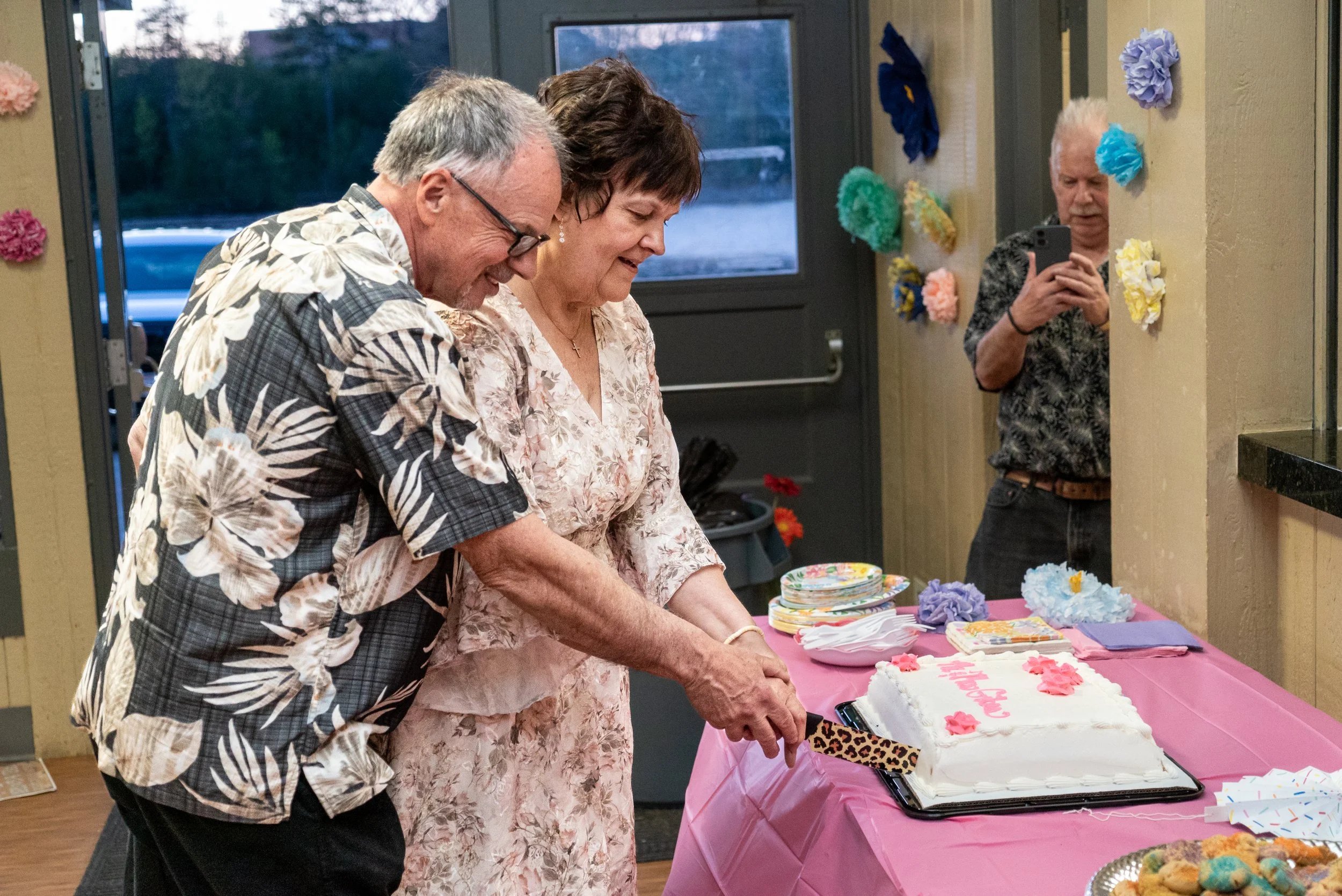 People cutting a cake at a celebration, with colorful decorations on the wall and a pink tablecloth on the table.