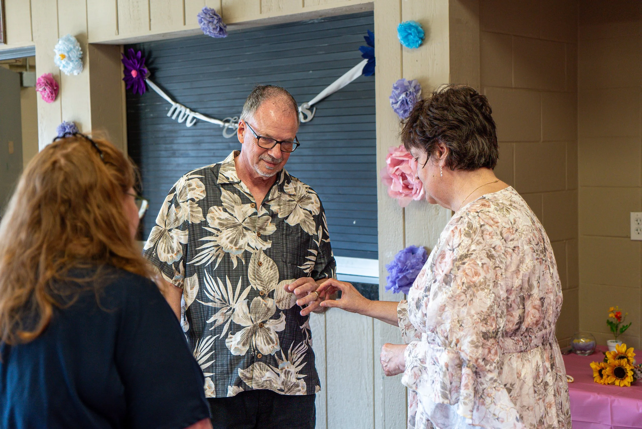 A couple exchanging rings at a casual outdoor wedding ceremony with decorated walls and a table with sunflowers.