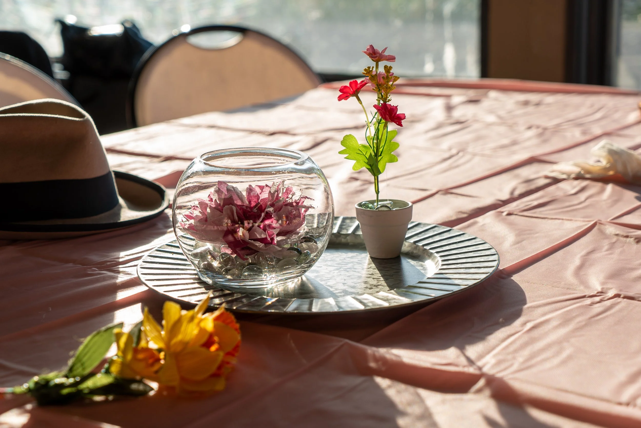 A table with a pink tablecloth set with decorative items including a small flower arrangement in a white pot, a glass bowl filled with pink flowers and decorative stones, and a straw hat with a black band. Sunlight streams in through a window in the 