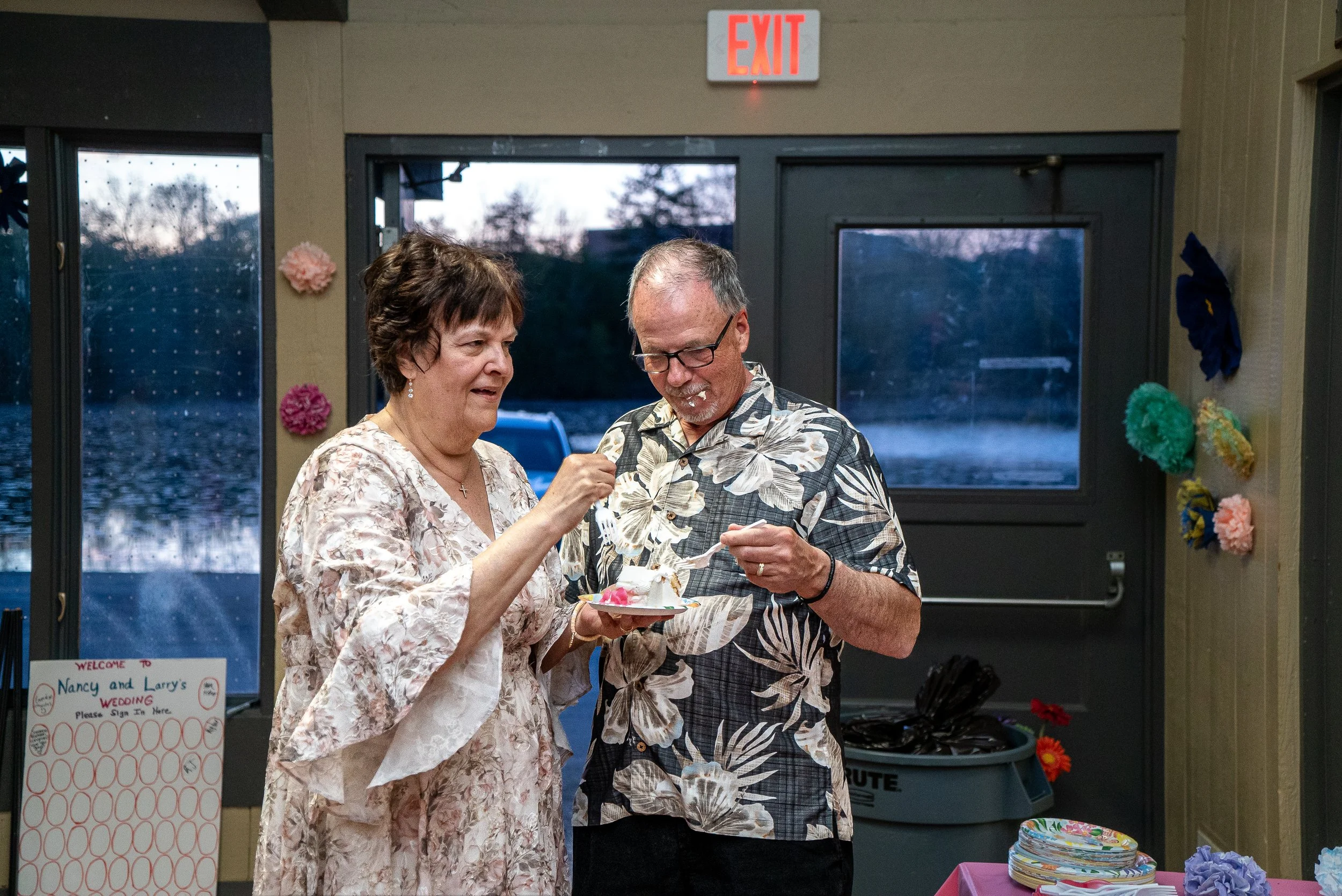 A woman and a man are at a celebration, possibly a wedding reception, with the woman holding a plate of cake and the man taking a piece; they are indoors near a doorway with a red exit sign above. Decorations and signs related to Nancy and Larry's we