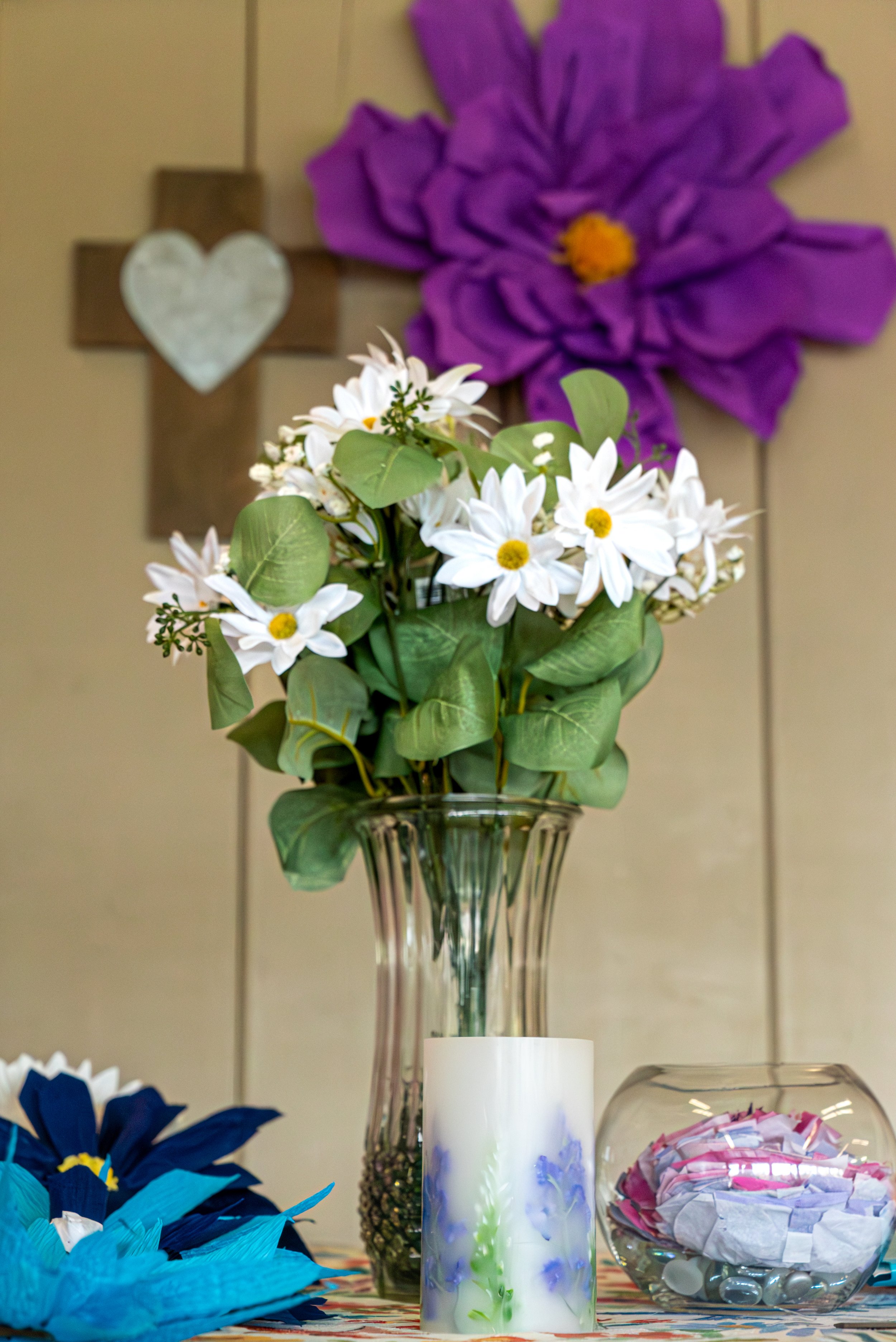 Decorative floral wedding arrangement featuring white daisies with yellow centers and green leaves in a clear glass vase, a white and blue marbled candle, a large purple paper flower, and a bowl filled with shredded colored paper and decorative stone