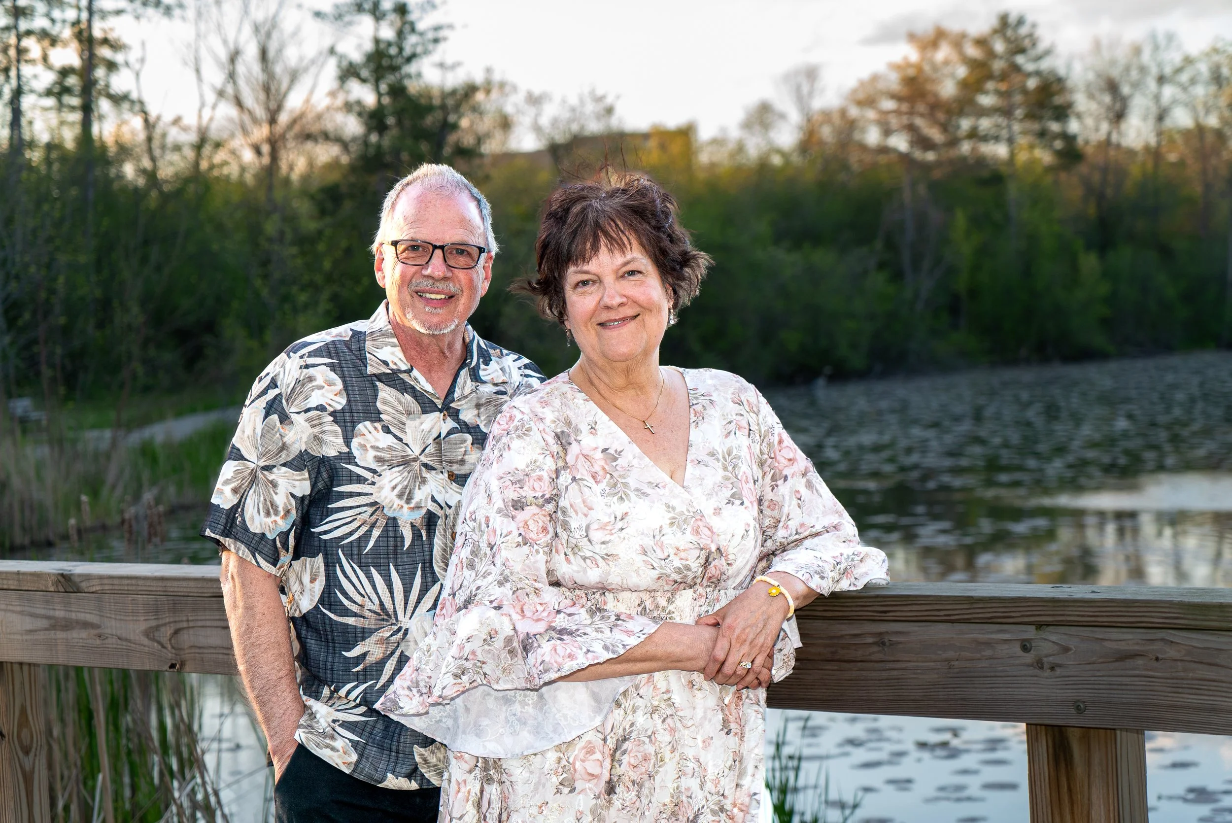 A smiling older man and woman standing outdoors on a wooden deck near a body of water with trees and a house in the background during late afternoon or early evening.
