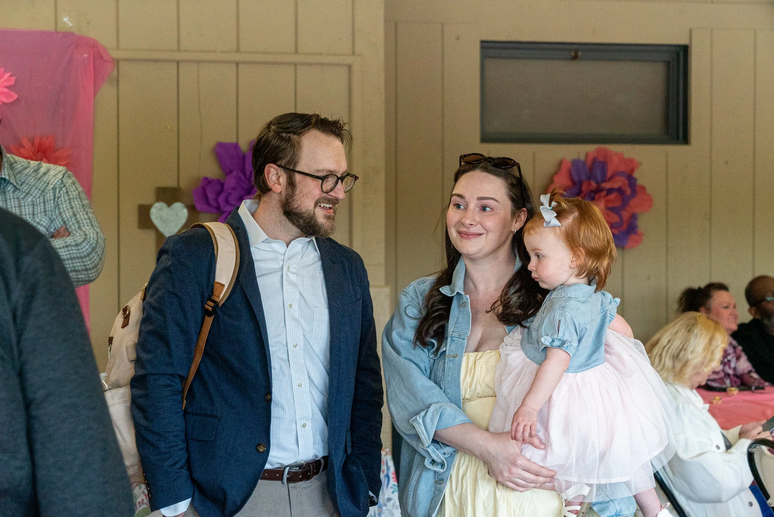 A man, woman, and young girl are at a wedding, engaging in conversation. The man, with glasses and a beard, wears a dark blazer and a light shirt. The woman has long brown hair, sunglasses on her head, and wears a denim jacket. The girl has red hair,