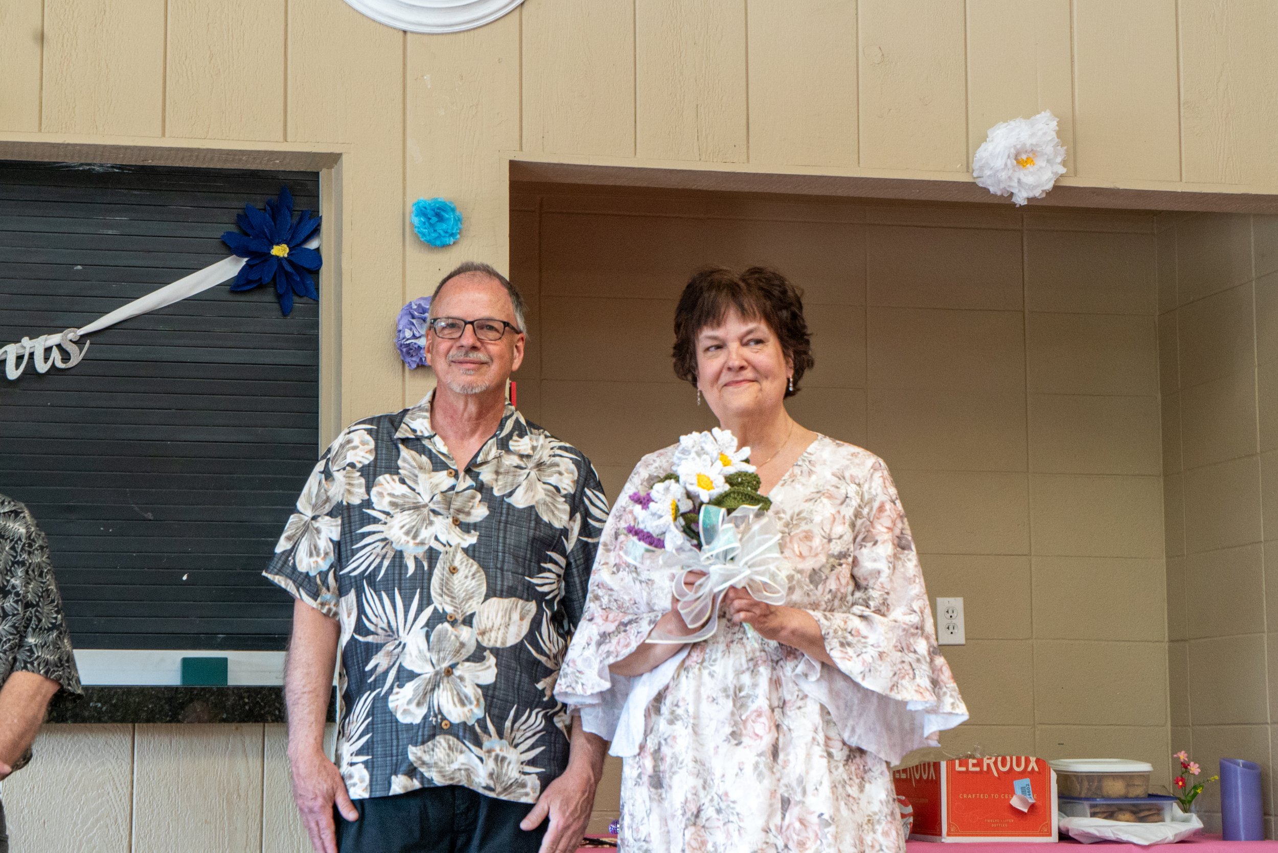 A man and a woman standing side by side indoors, the woman holding a bouquet of flowers. Both are dressed in patterned shirts. The background features decorated walls with paper flowers and a banner.