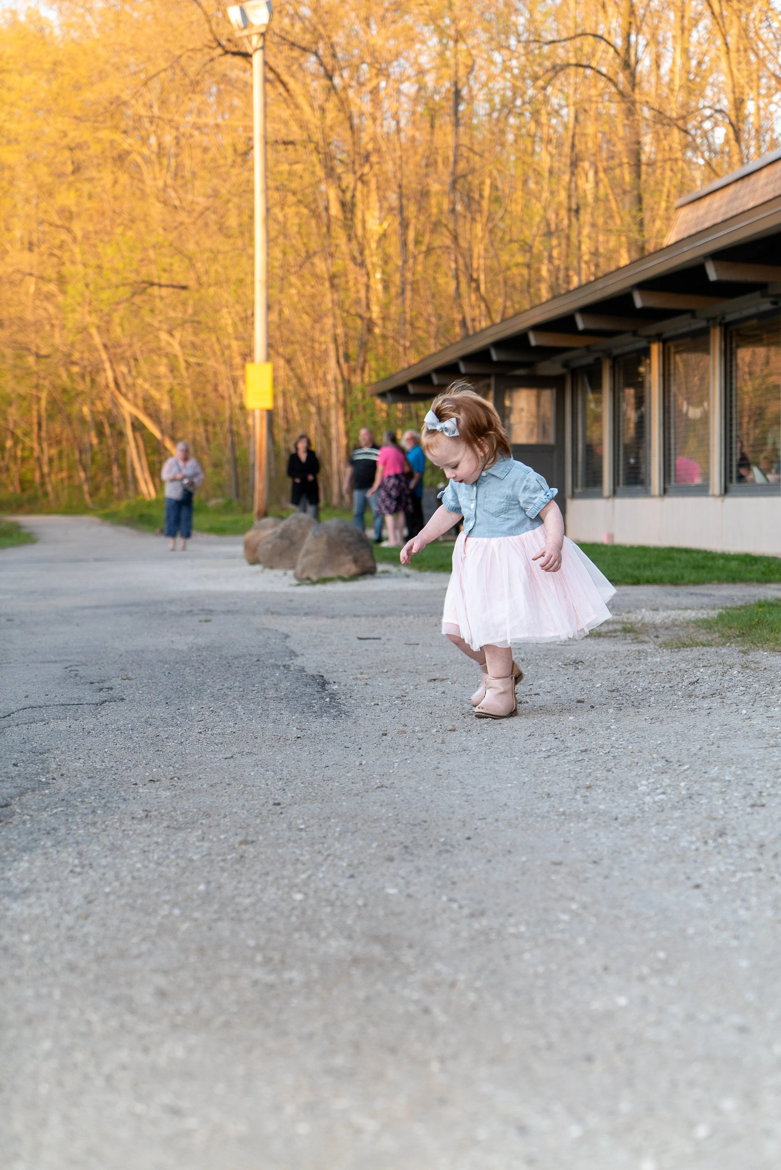 A young girl with a bow in her hair is walking on a gravel area, wearing a light blue top and a pink tulle skirt. In the background, several people are standing near a building, with trees and a pole visible.