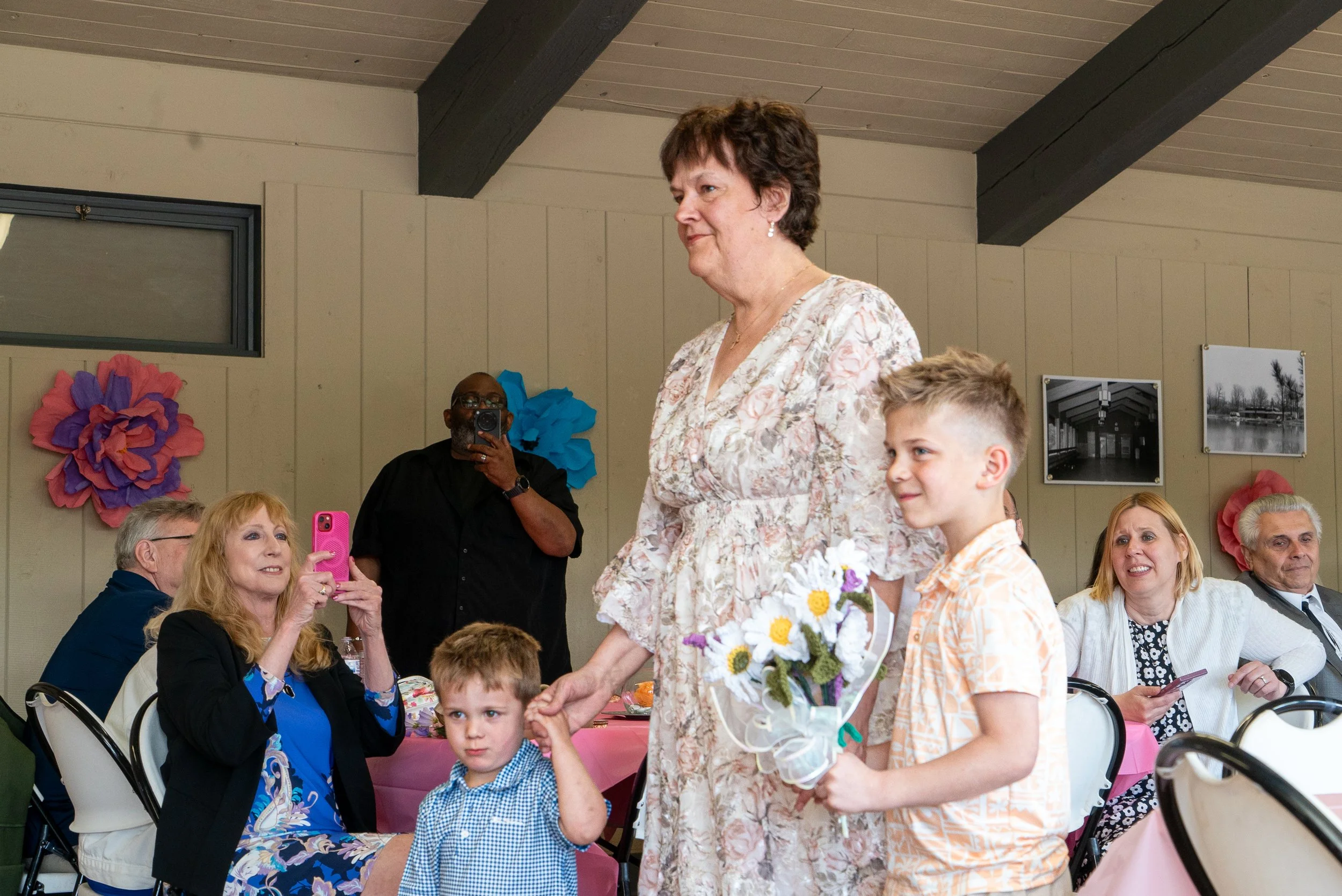 A woman wearing a floral dress is presenting a bouquet of flowers to a boy holding a floral arrangement at a gathering. Multiple people are seated around a pink table, some taking photos, in a decorated room with large paper flowers and framed photos