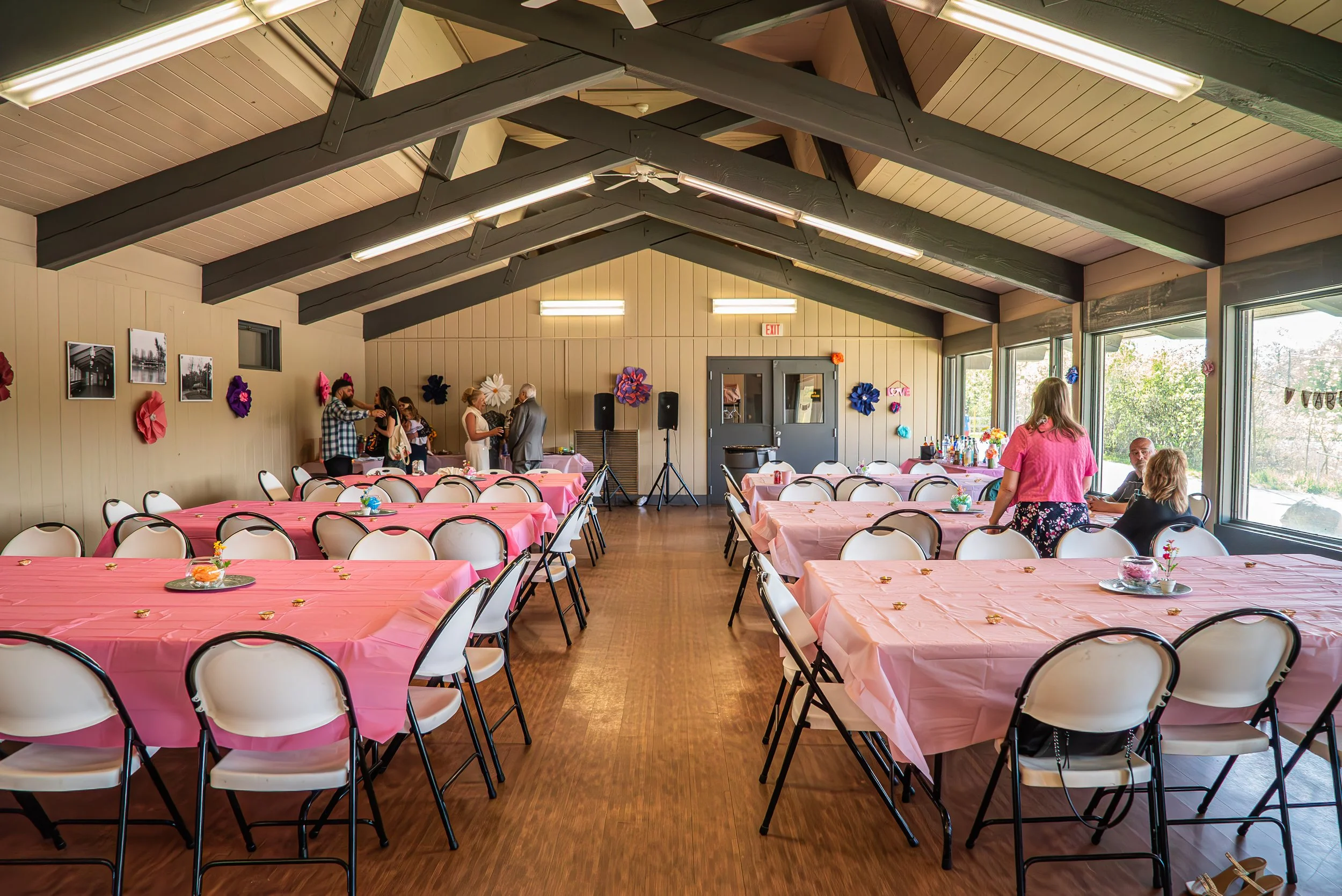 Decorated banquet hall with pink tablecloths, folding chairs, wall art, and flower decorations, with people mingling by a bar area before the wedding.