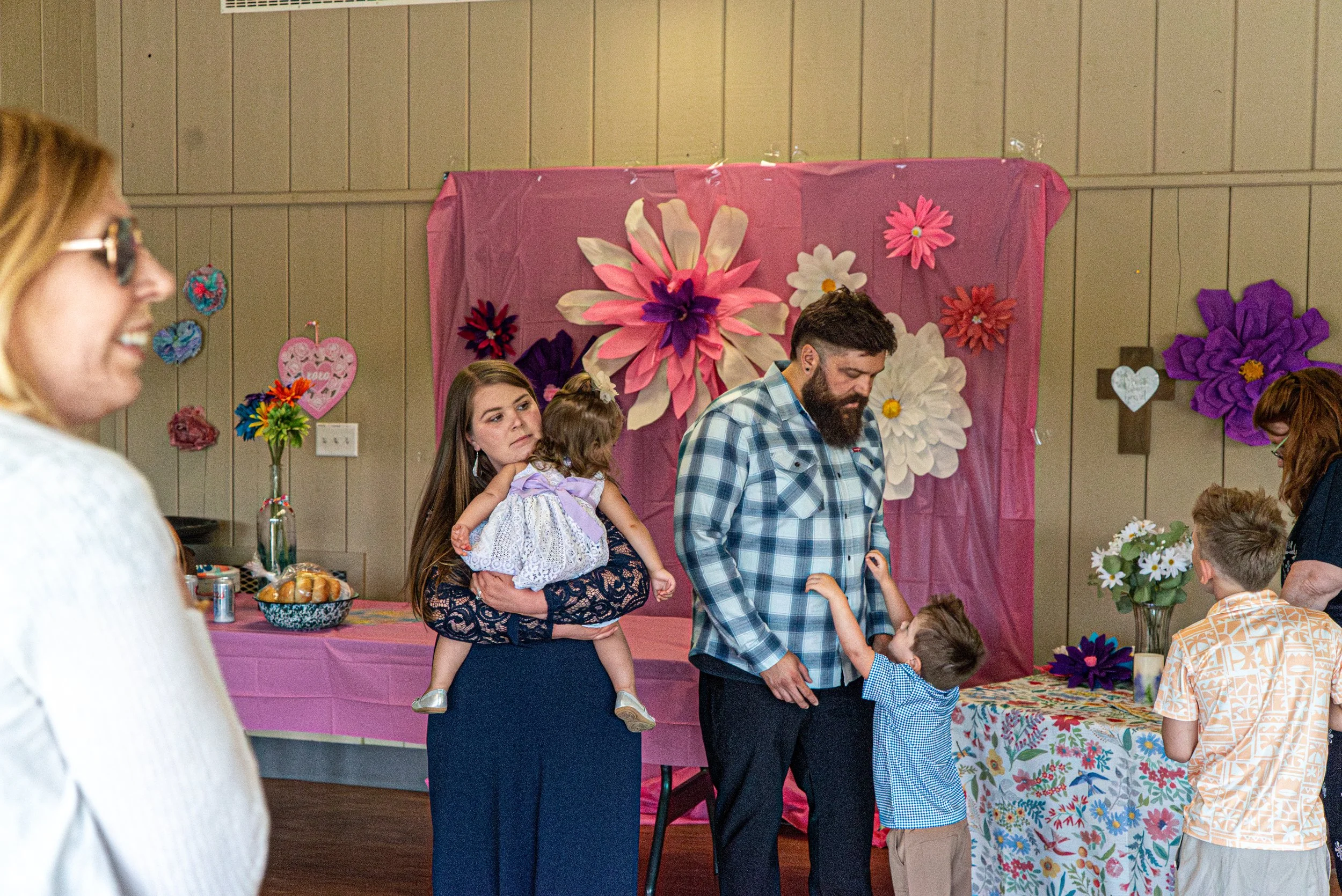 A woman is holding a young girl on her hip in a room decorated for a celebration with large paper flowers and heart-shaped ornaments on the walls. Other people are present, including a man shaking hands with a boy, a woman smiling, and a person weari