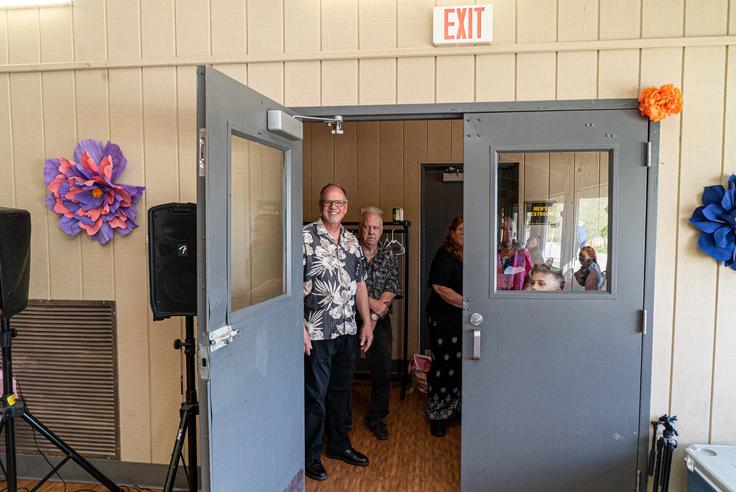 People gathering at the entrance of a building with decorative flowers on the wall, an exit sign above, and a window reflecting other people inside.