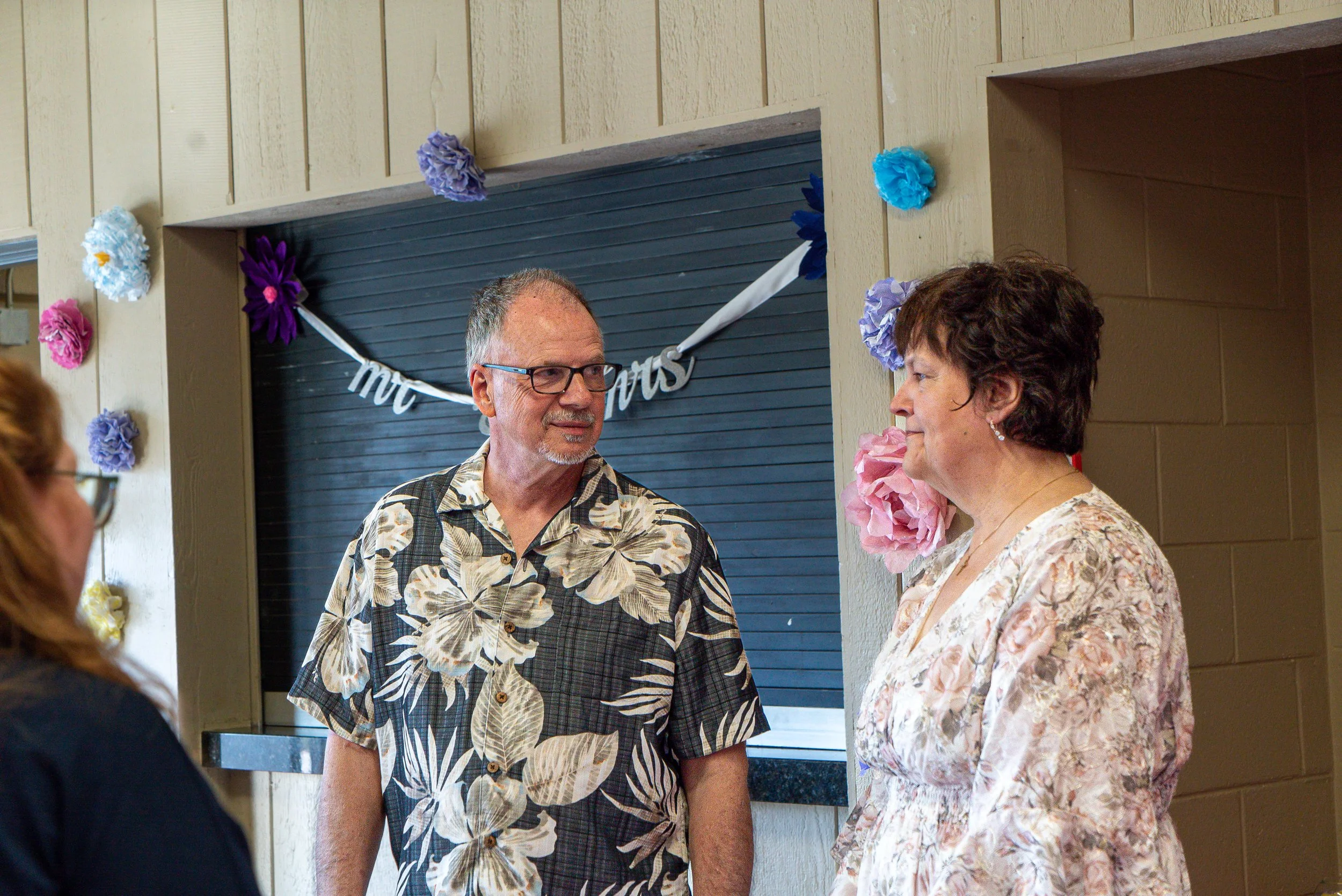 A man wearing glasses and a floral shirt talking to a woman with short brown hair wearing a floral blouse at an indoor event decorated with colorful paper flowers and a "Mr. & Mrs." banner.