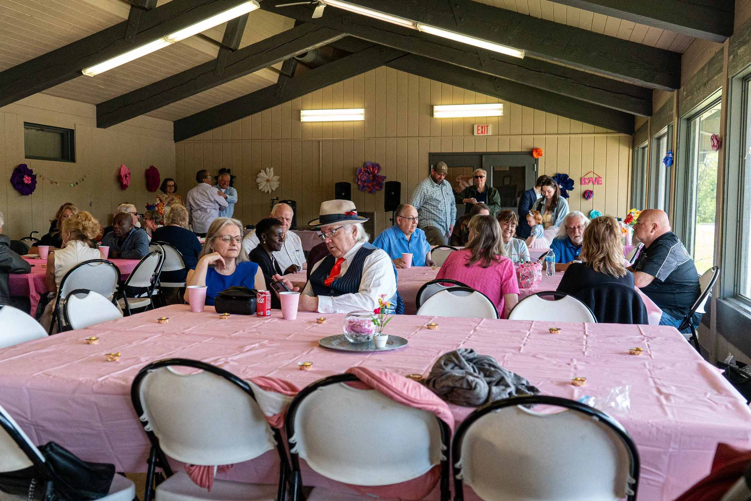 People gathered in a decorated indoor wedding venue with pink tablecloths, large paper flowers on the walls, and sunlight coming through large windows.