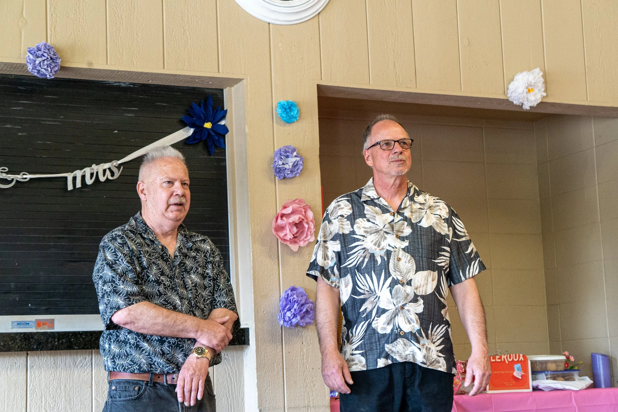 Two men are standing indoors at a celebration, wearing tropical shirts. The background is decorated with colorful paper flowers and a blackboard with some unclear writing, indicating a festive or special occasion.