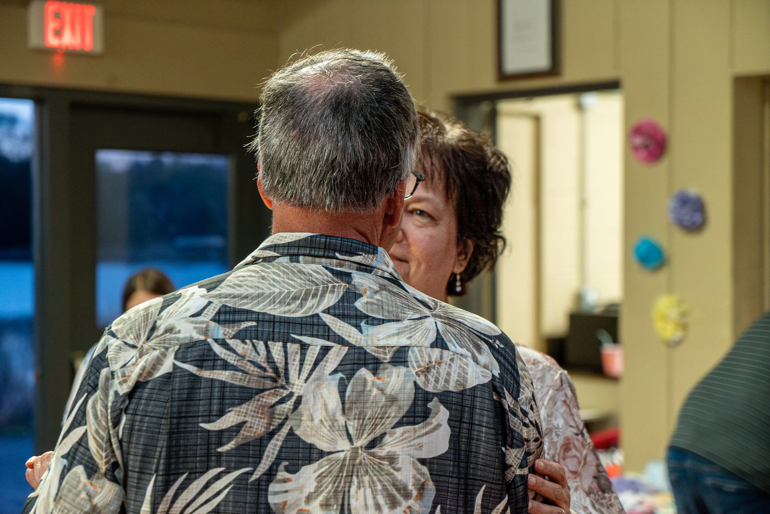 A middle-aged man and woman are hugging warmly indoors, with a blurred background showing a door, wall decorations, and a table. The man has gray hair and is wearing glasses and a patterned shirt, while the woman has dark hair and is wearing earrings
