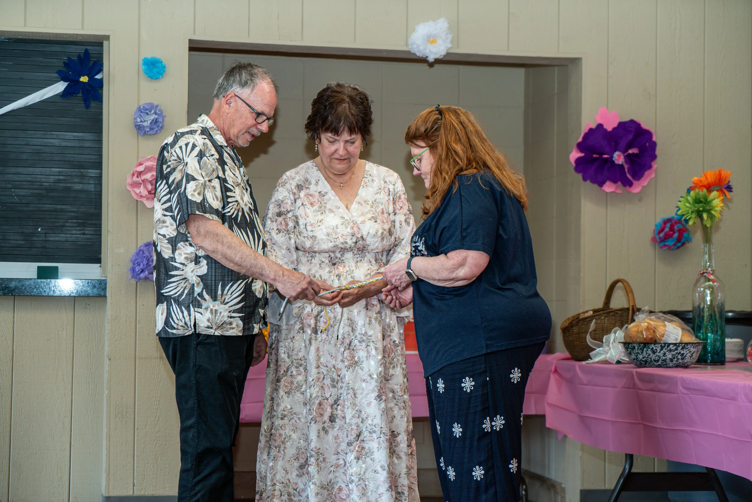Three people standing closely together, looking at a small object or paper one person is holding. They are in a decorated room with colorful paper flowers on the wall and a table with pink tablecloths and bowls of snacks.