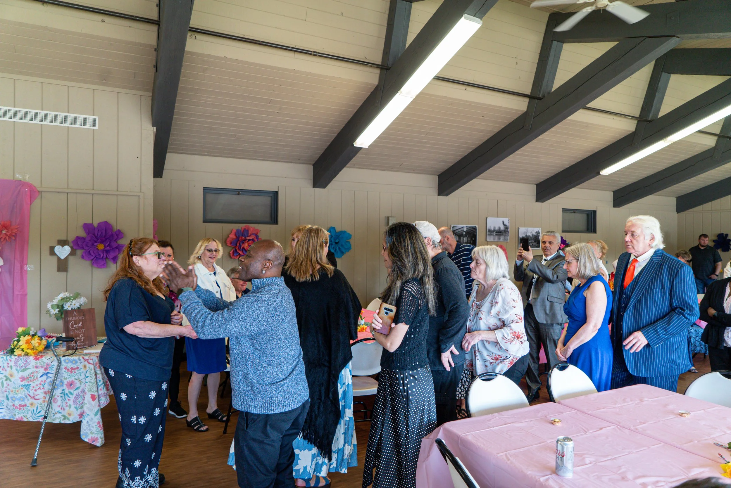 People gathered at a social event in a decorated room, chatting and taking photos near tables with pink tablecloths and floral decorations.