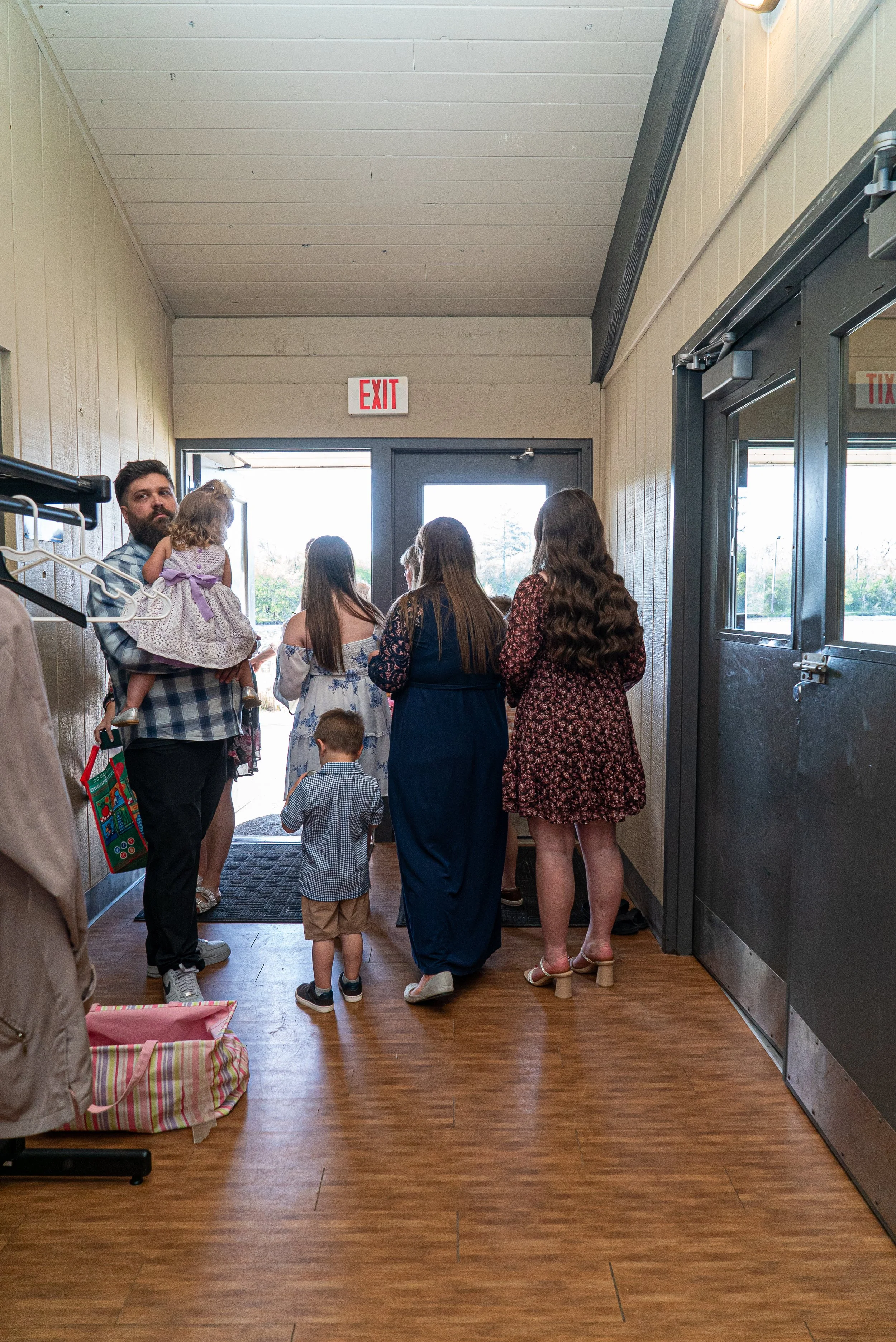 People standing in line right inside an entrance with a door at the back, wooden floors, and beige walls, some wearing casual summer clothing, including a man holding a small girl, and children among the group.