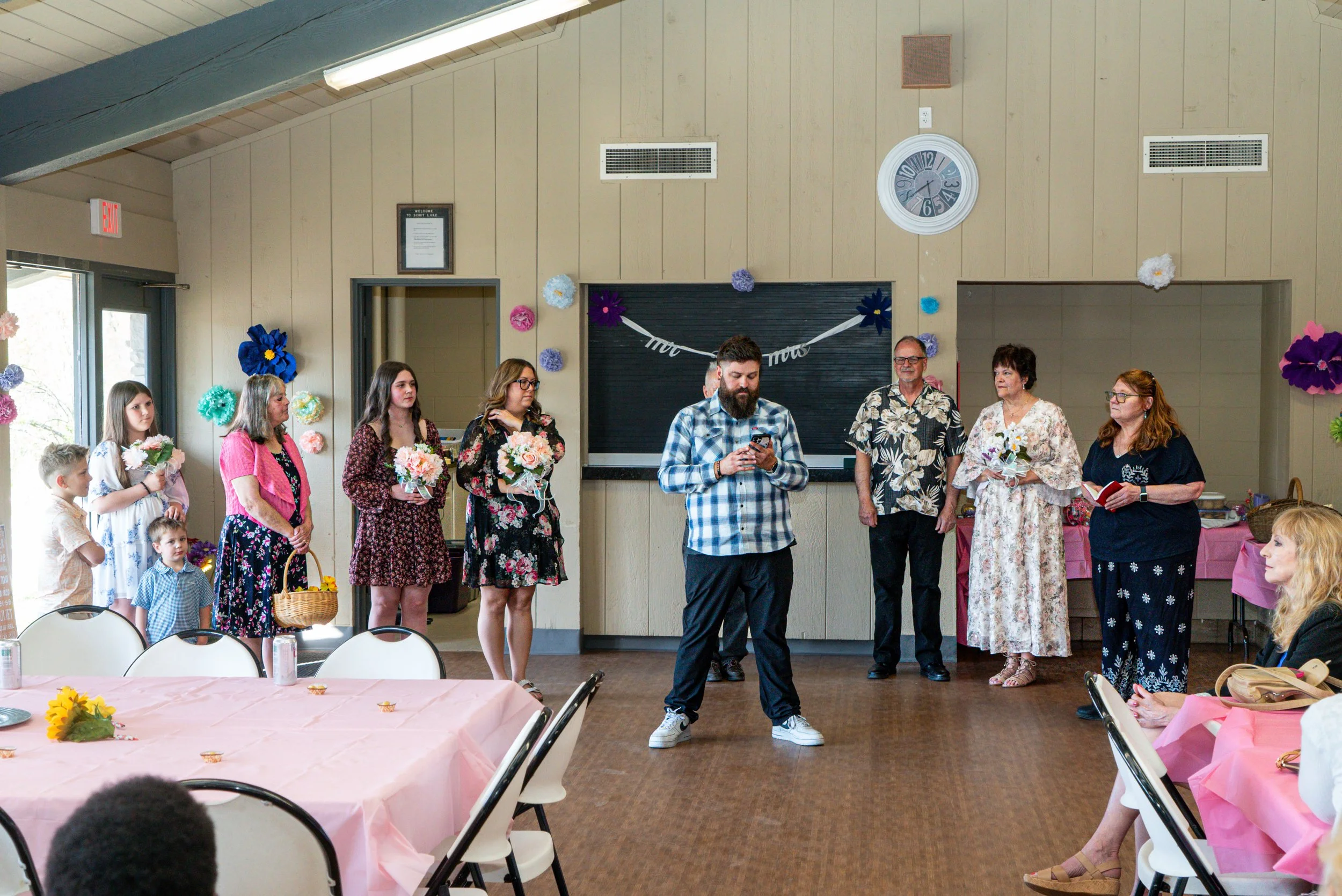 A group of people, including children and adults, standing in a decorated room with tables and chairs, at what appears to be a celebration or gathering. Some individuals hold flowers, and there are colorful paper flowers on the wall.