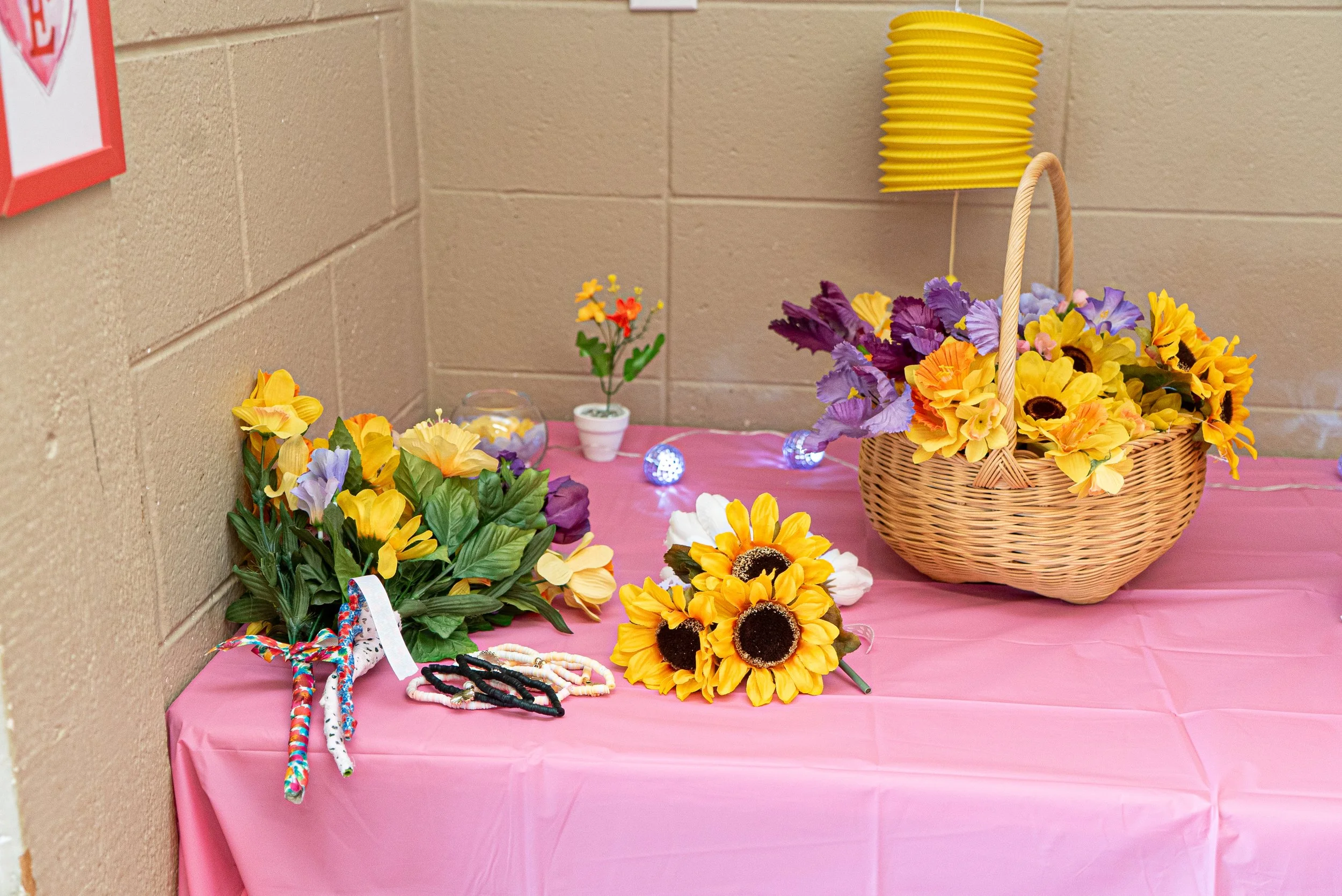 Display of colorful artificial sunflowers, purple and yellow flowers in a wicker basket, small decorative flowers, and jewelry on a pink tablecloth against a beige brick wall for a wedding.