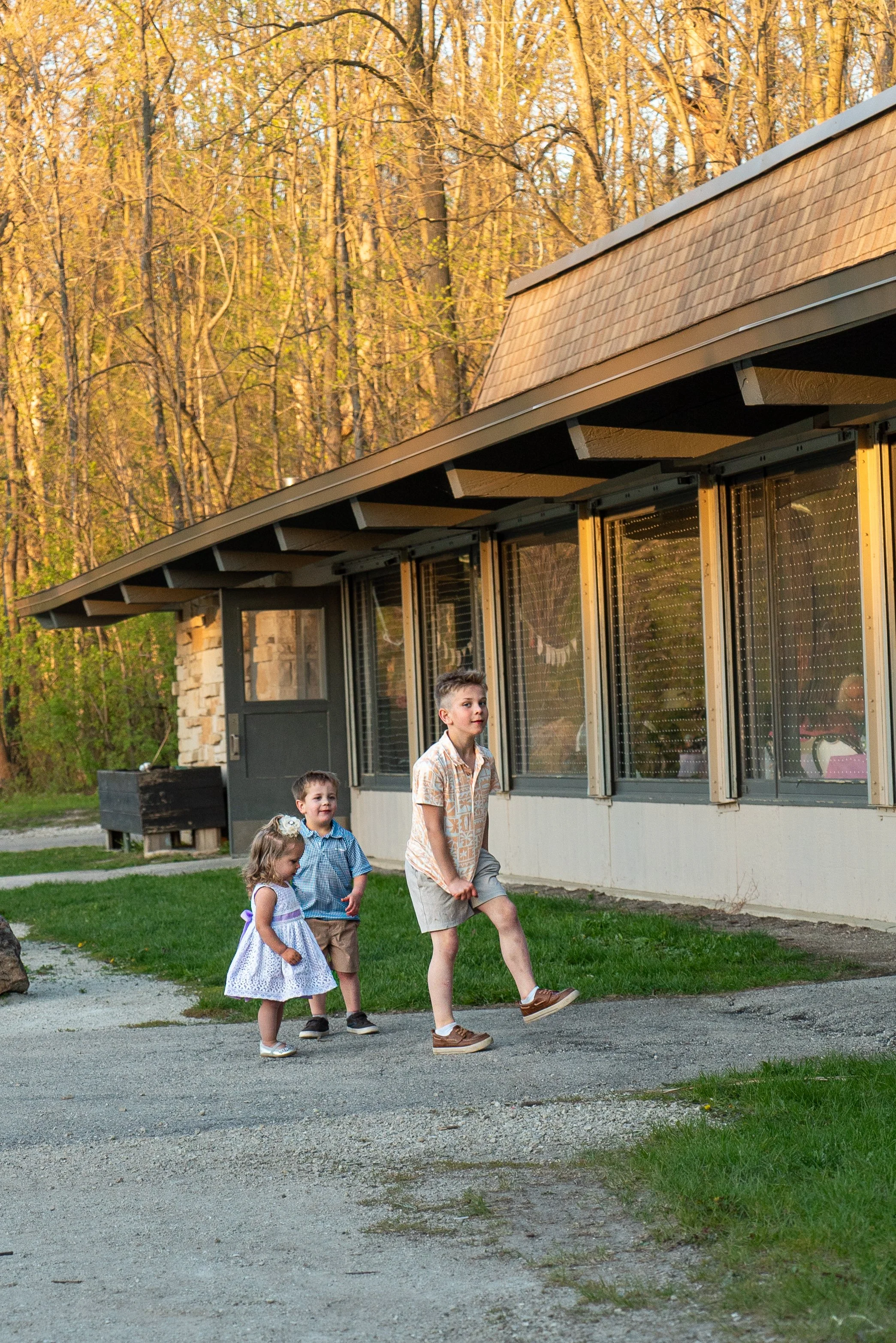 Four children walk outdoors near a building with large windows, set against a background of trees with fall foliage. The children include a girl in a white dress, a boy in a blue shirt, and two other boys in casual attire.