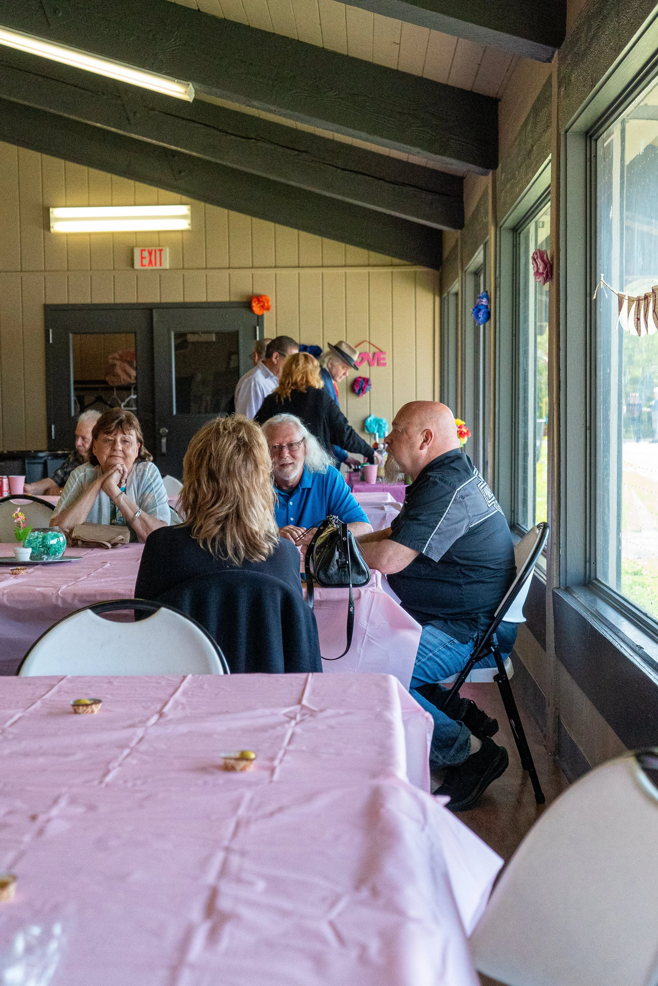 People sitting around a banquet table with pink tablecloths, inside a decorated room with large windows and a wood-paneled ceiling. Some people are talking, and others are preparing food or drinks. Decorative flowers and a banner are visible in the b