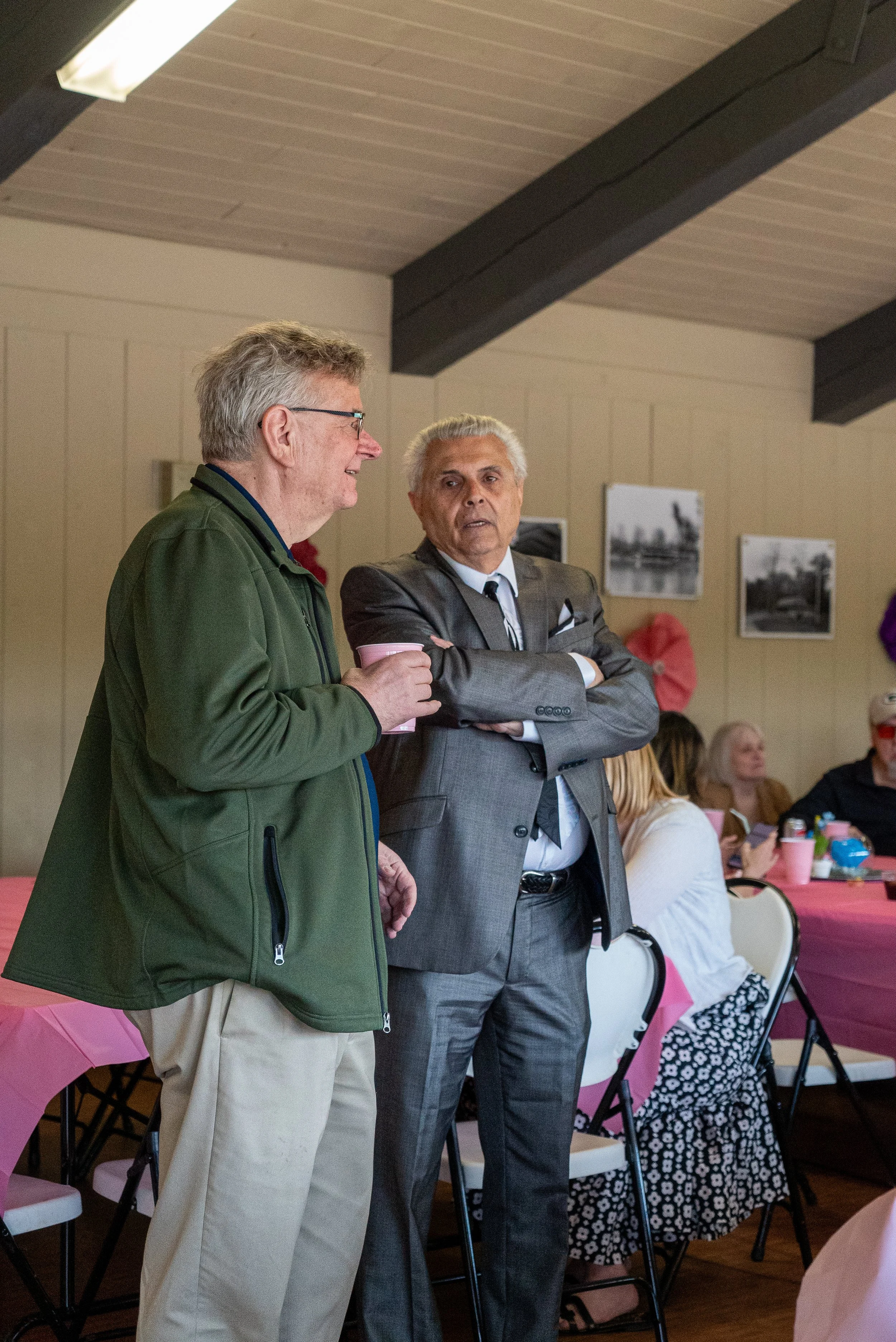 Two men standing and talking at a wedding, with several women sitting at tables in the background. The setting appears to be a banquet or event hall with wood-paneled walls and wall art.