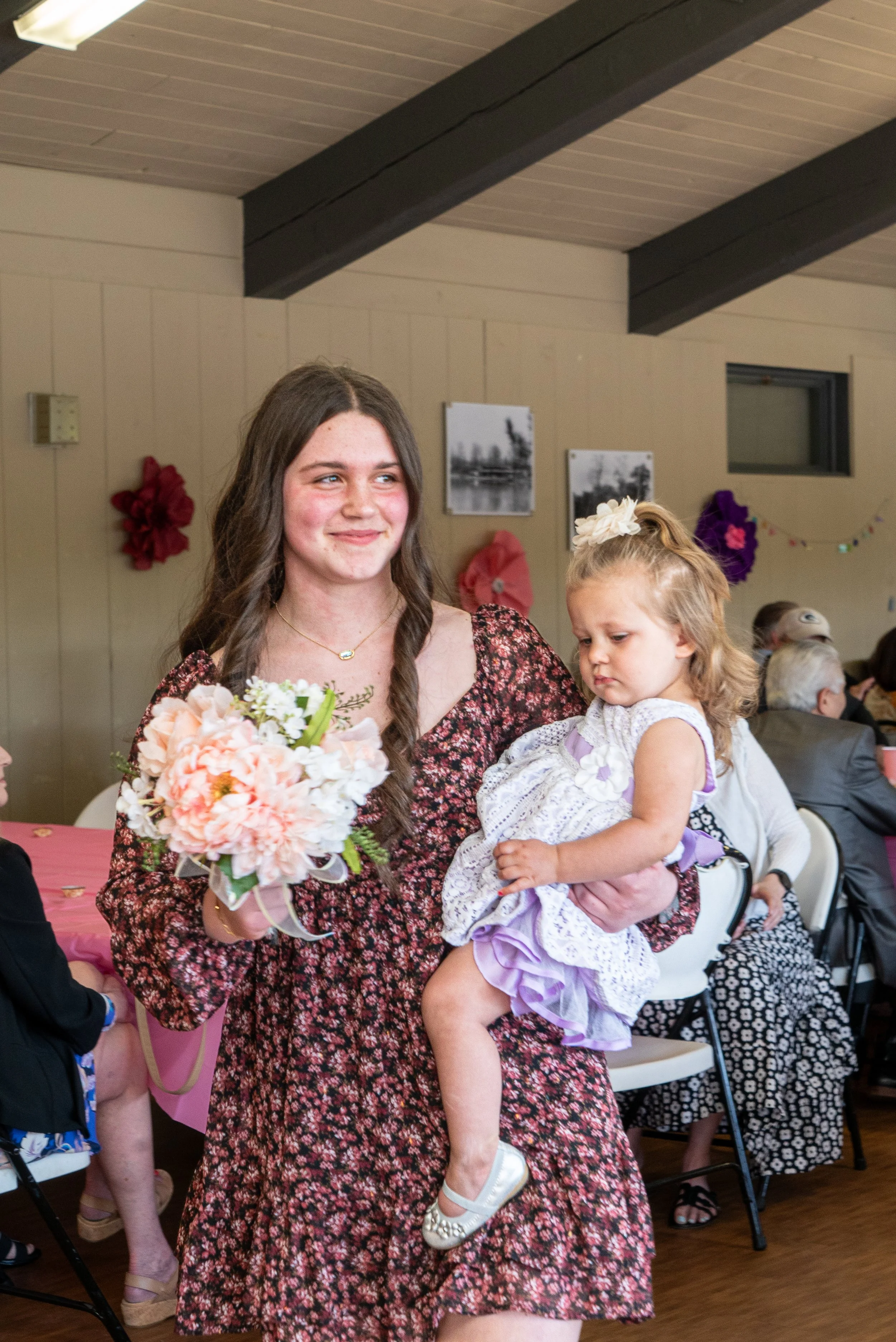 A woman with long brown hair in a floral dress holding a bouquet of flowers and a young girl dressed in a white and purple dress, with a flower headband, sitting on her arm at a celebration or gathering.
