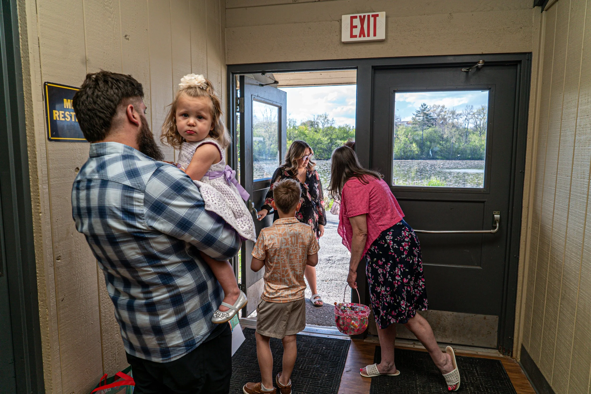 A group of people, including children and adults, are entering or exiting through a door with a window leading outdoors, where trees and a lake are visible.