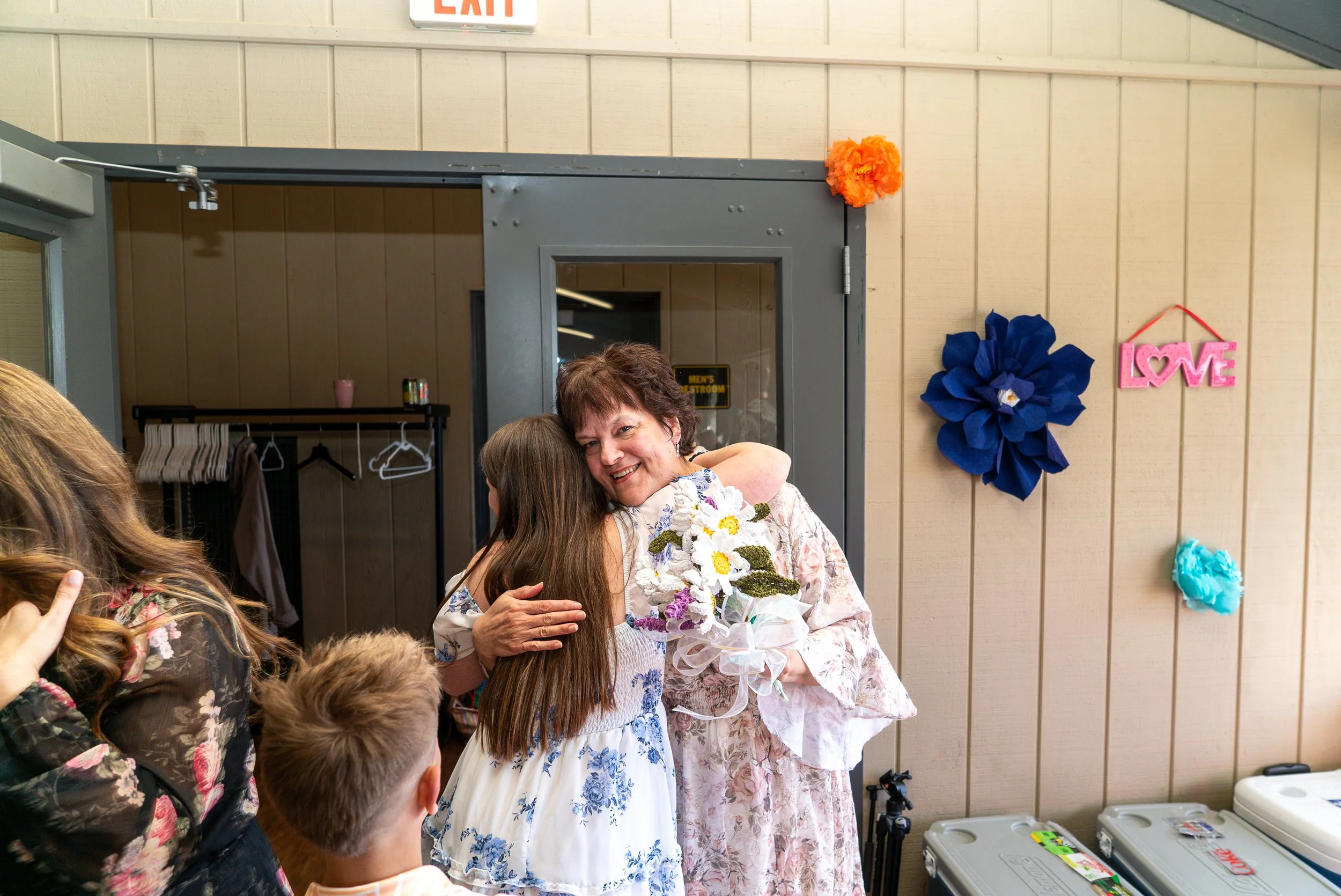 A woman hugging a girl holding a bouquet of flowers during a celebration with people gathered around. Decorations include bright orange and blue paper flowers, and a pink "Love" sign on the wall. An exit sign is visible above the door.