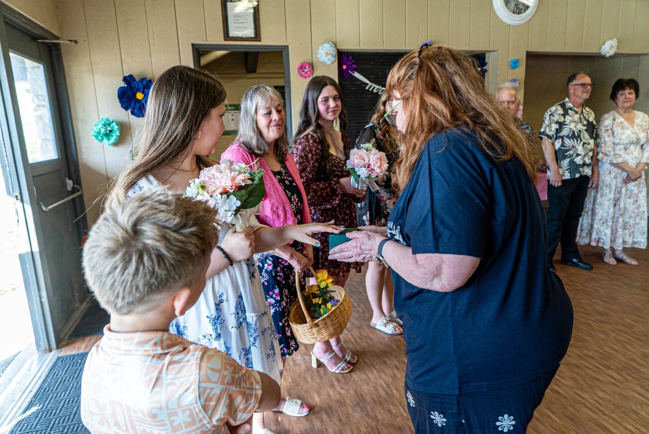 A woman with red hair and glasses is handing a small gift to a young girl at a celebration. The girl is holding flowers and a basket, and is accompanied by other women and children, some holding flowers. The setting is decorated with colorful paper f