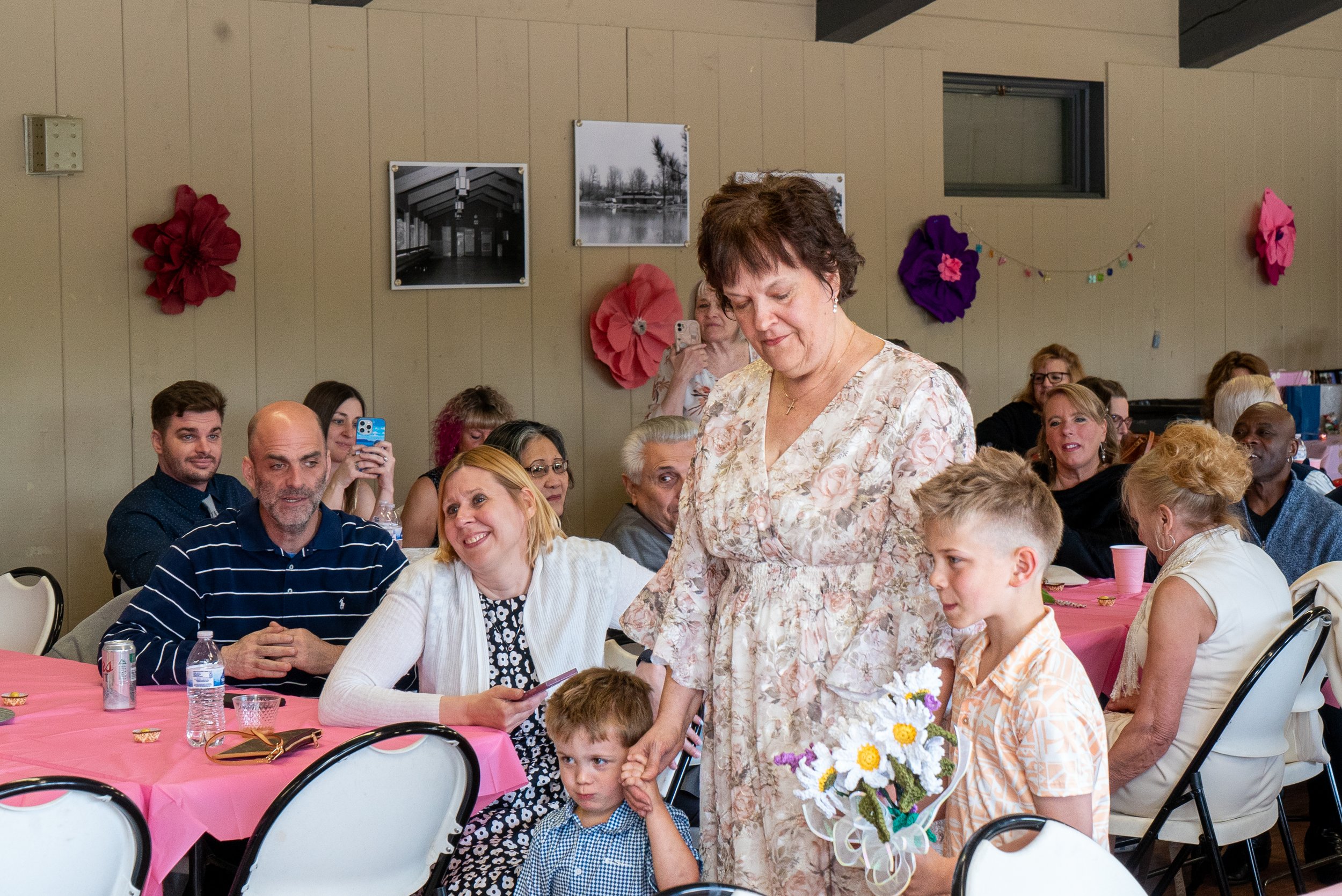 A group of people sitting at pink tablecloth-covered tables inside a room decorated with large paper flowers on the beige wall. Two women and two children are at the forefront, with a woman in a floral dress standing, a woman in a white sweater, and 