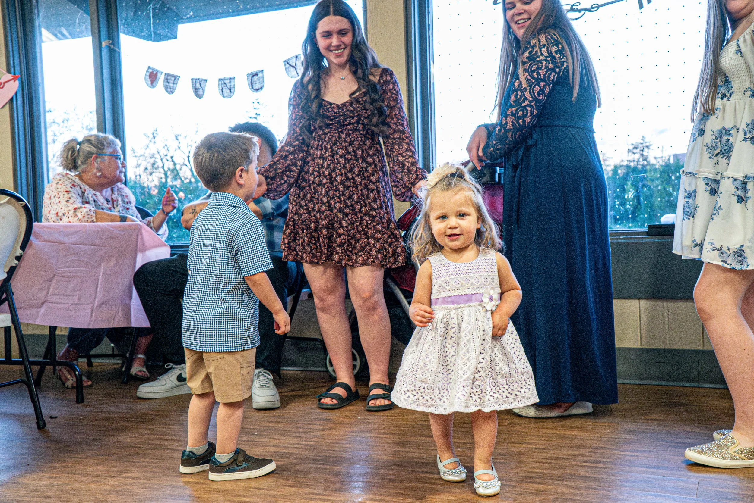 Children and adults at a small gathering or party, with some children standing and one girl in a white dress and two women dressed in darker clothes, inside a room with large windows and wood flooring.