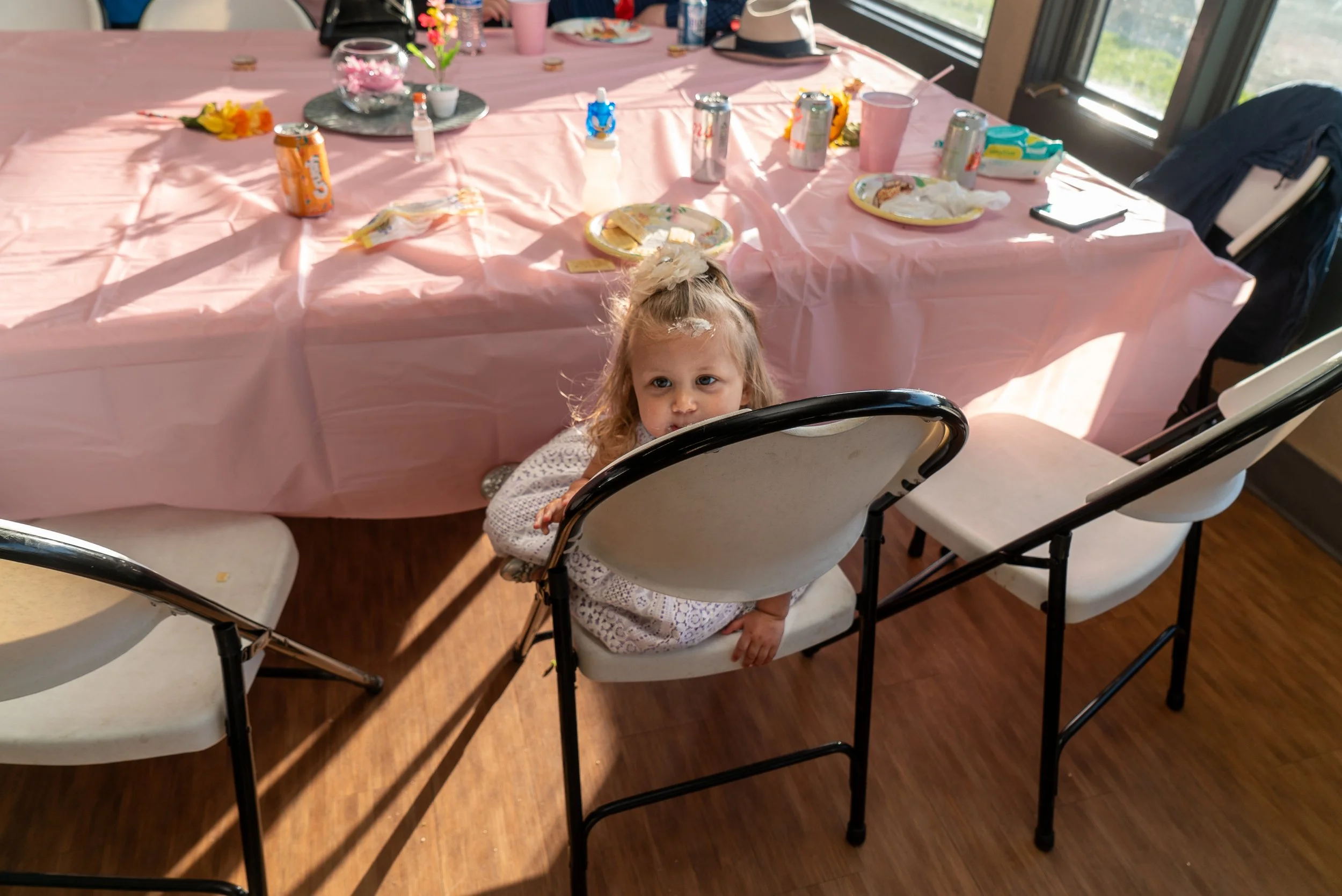 A young girl with curly hair and a floral hair accessory sitting in a high chair at a table. The table has a pink tablecloth and is set with plates, cups, soda cans, and partially eaten food. The table is next to large windows letting in natural ligh