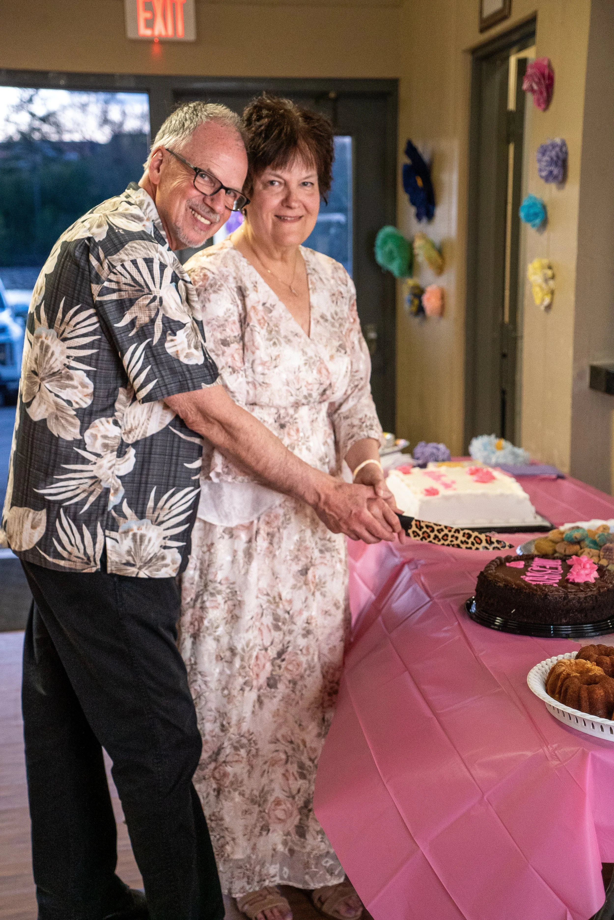 A man and woman smiling and holding hands at a celebration with a table of desserts, including a birthday cake with pink frosting.