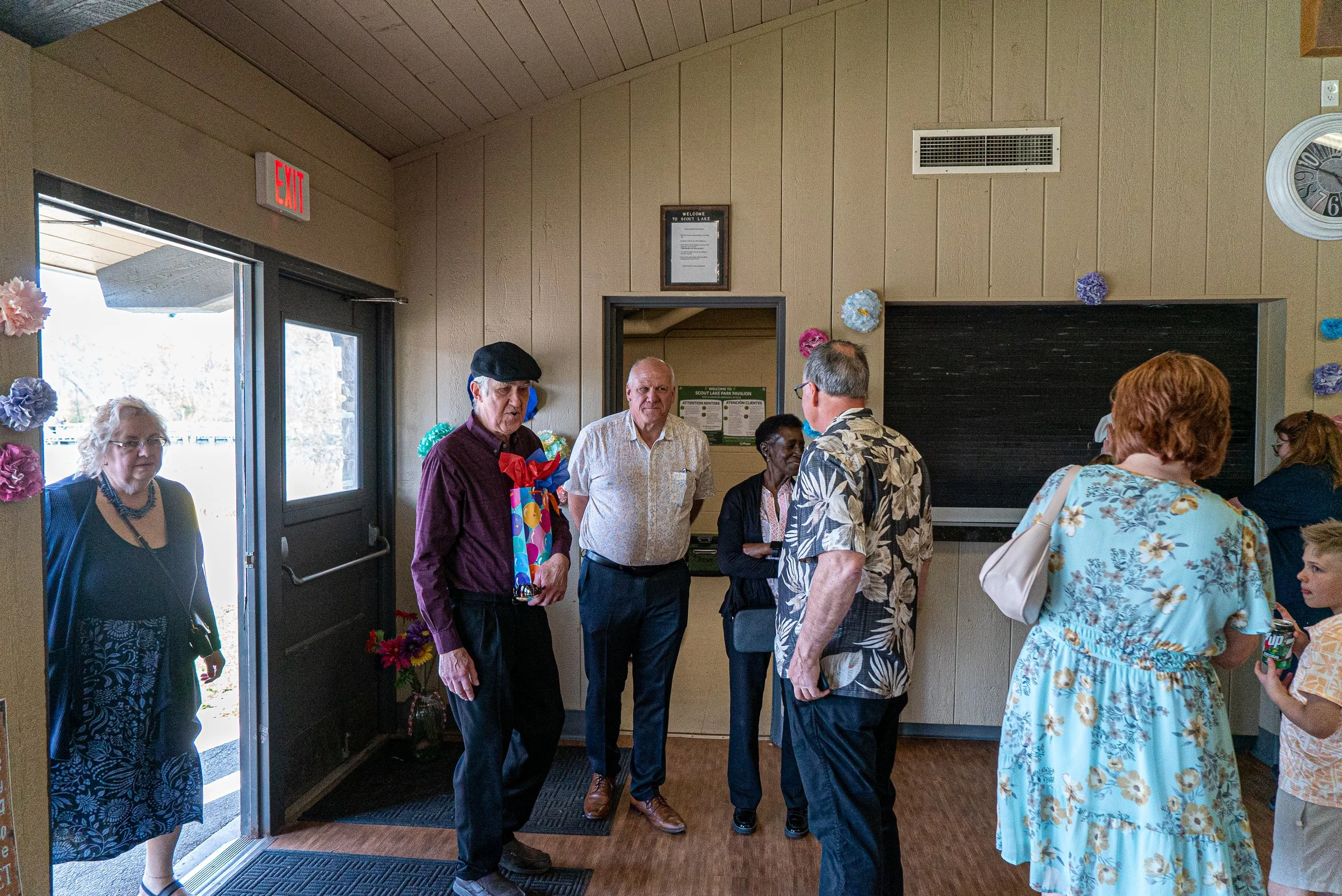 People gathered inside a building near the entrance, some engaged in conversation, with floral decorations on the wall.