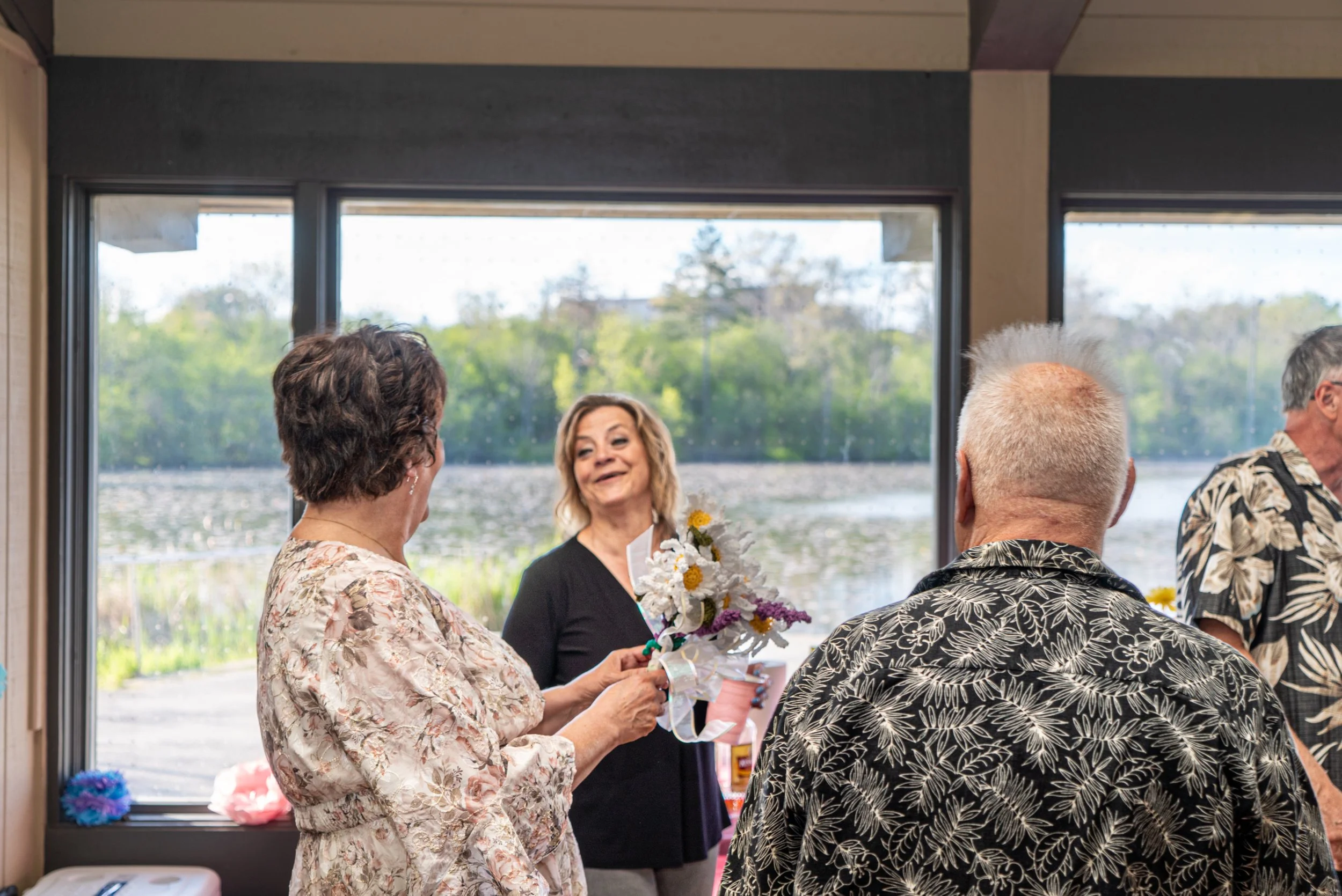 A group of people gathered indoors, with a woman holding a bouquet of flowers and smiling, engaging in conversation with others, near a window with a view of trees and water outside.