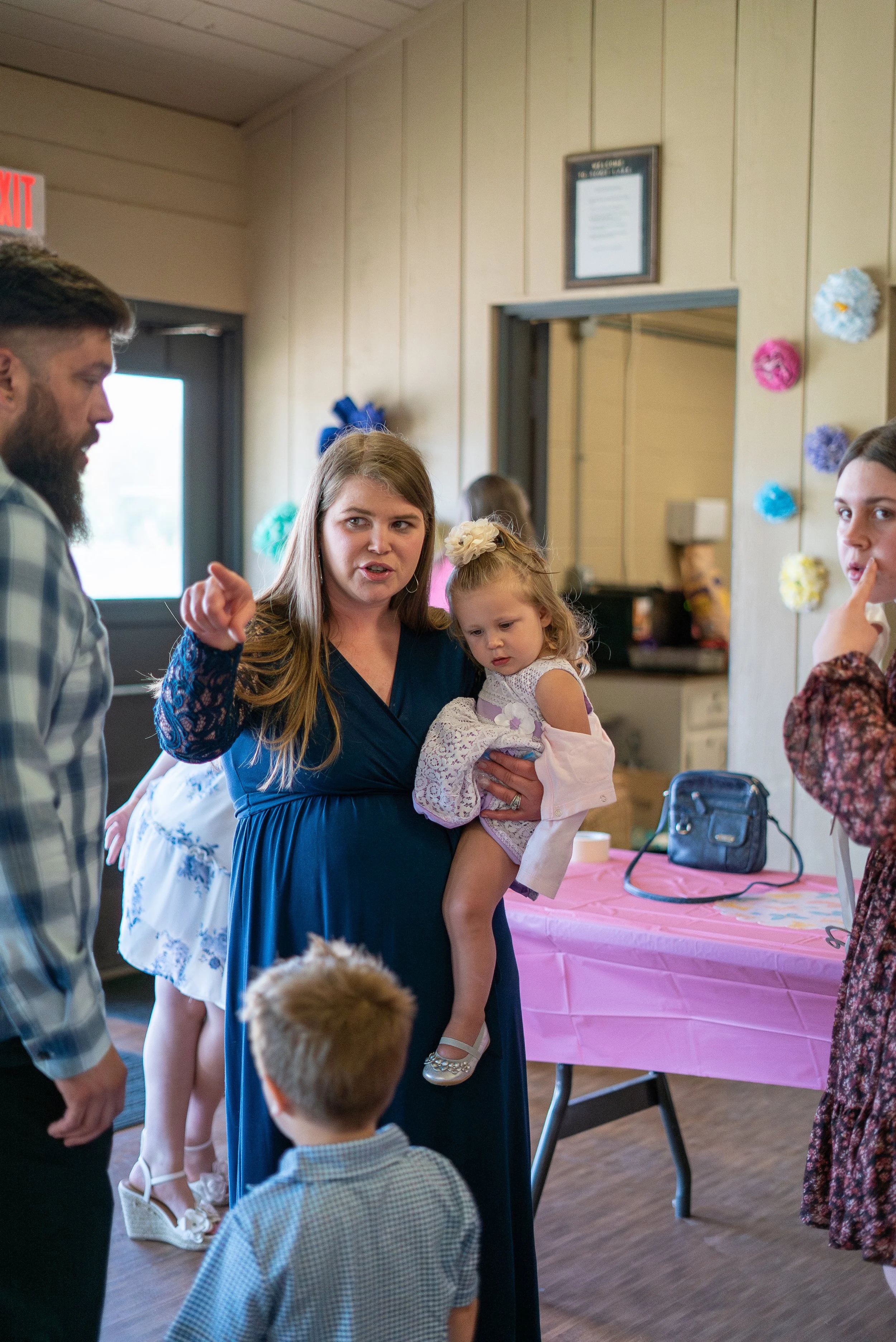 A woman holding a young girl with a flower headband, engaged in conversation with a man and another woman at a wedding with colorful decorations on the wall and a pink tablecloth.