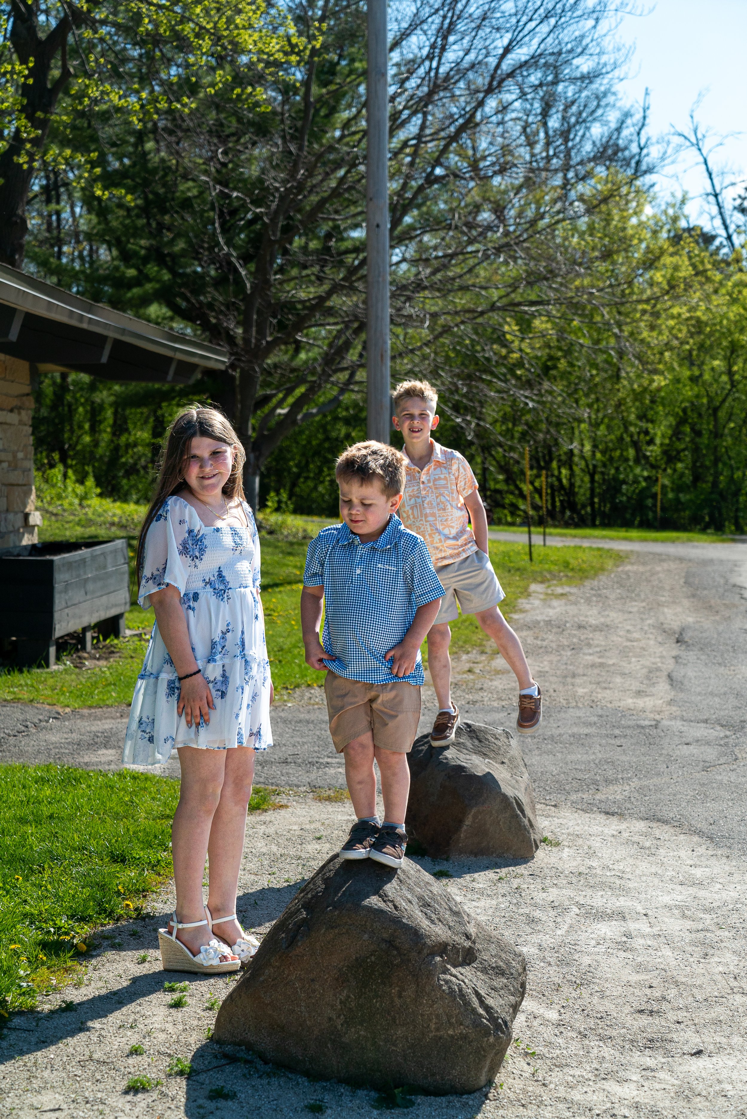 Three children are playing outside near large rocks on a sunny day. The girl in the foreground wears a white dress with blue floral patterns and white heels, smiling at the camera. Two boys stand in the background, one standing on a rock, both smilin