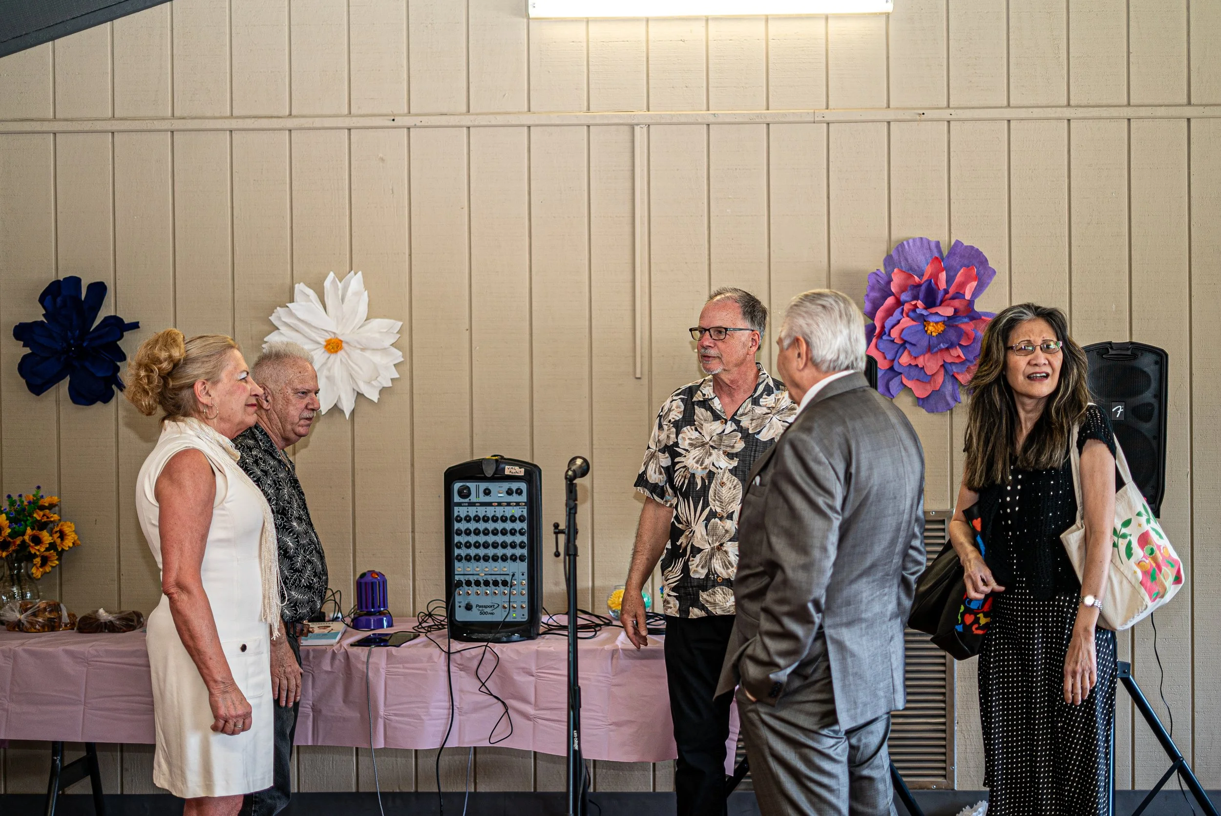 A group of people at a wedding, standing near a table with audio equipment, decorated with large paper flowers on a beige wall behind them.