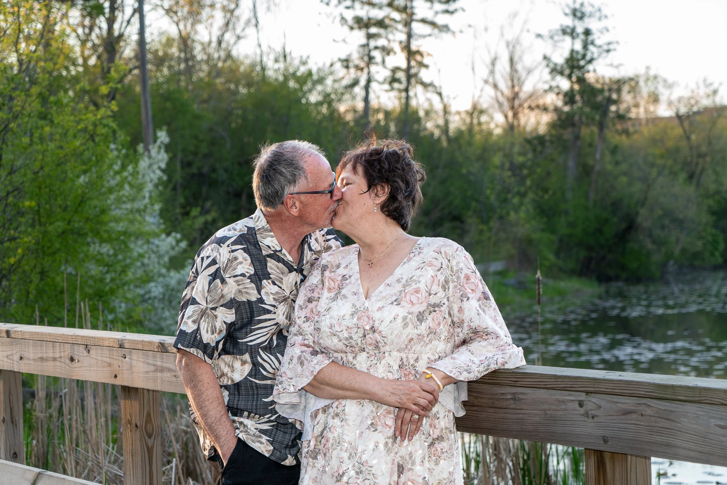 An elderly couple sharing a kiss by a lake, standing on a wooden dock with a green, forested background at sunset.
