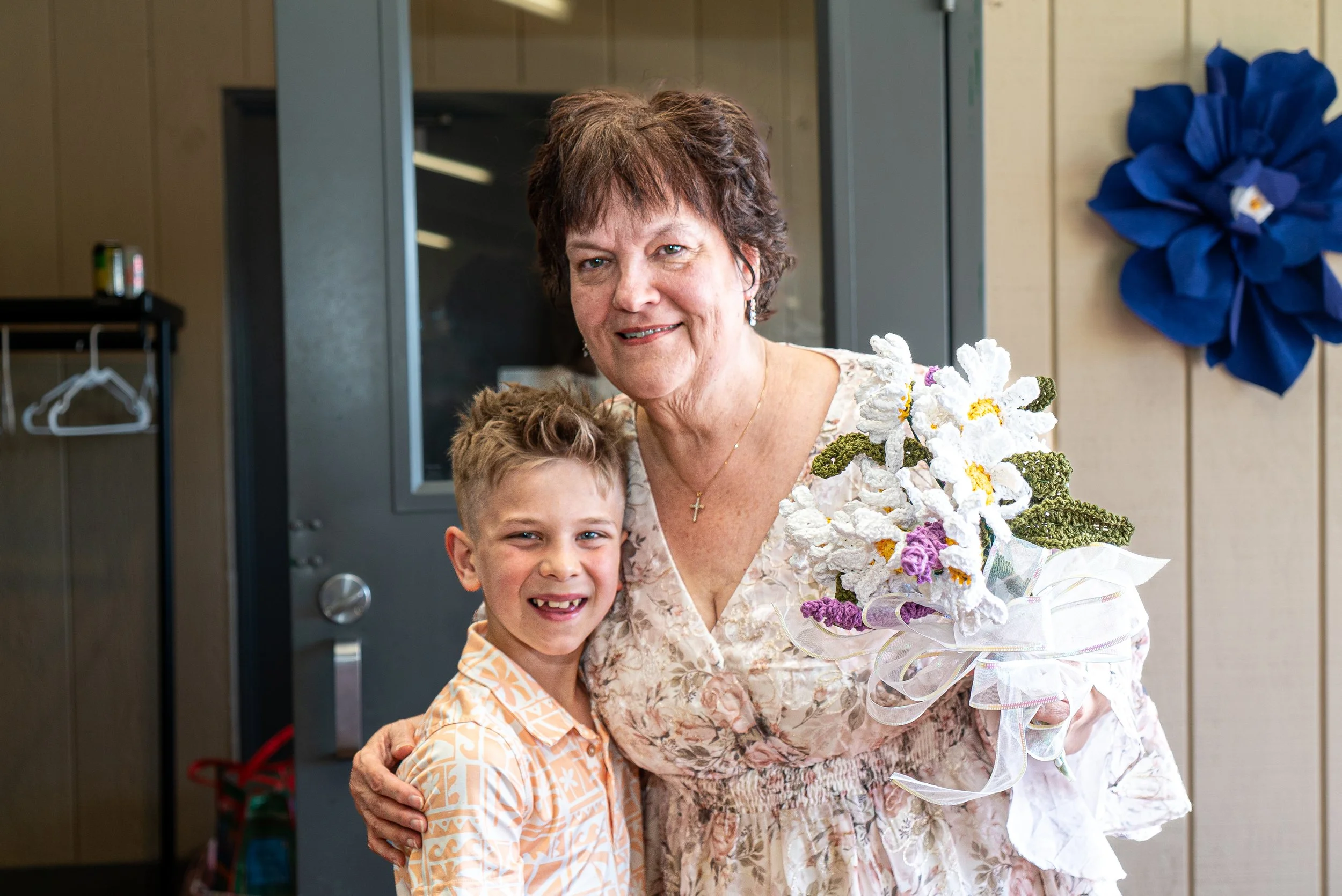 An elderly woman holding a bouquet of white and purple flowers, smiling, with a young boy in an orange patterned shirt standing beside her, both hugging in an indoor setting.