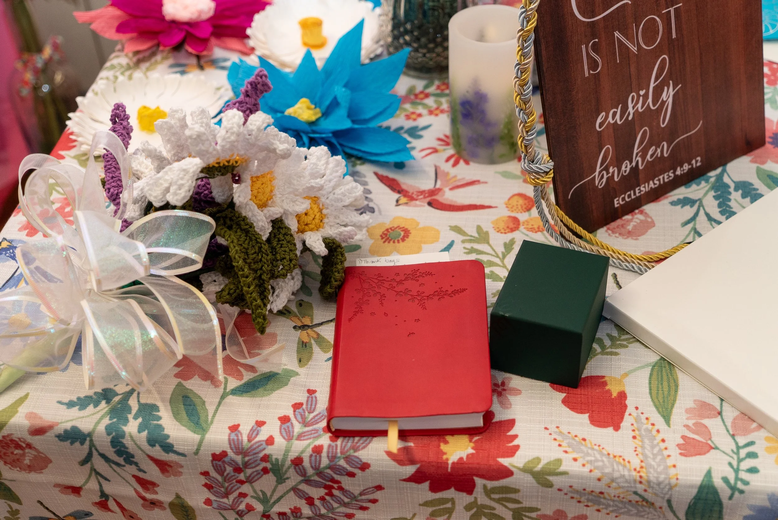 Decorative table setting with a floral tablecloth, crocheted flowers, a red notebook, a green box, and a sign that reads 'Easter is not a activity, broken'.