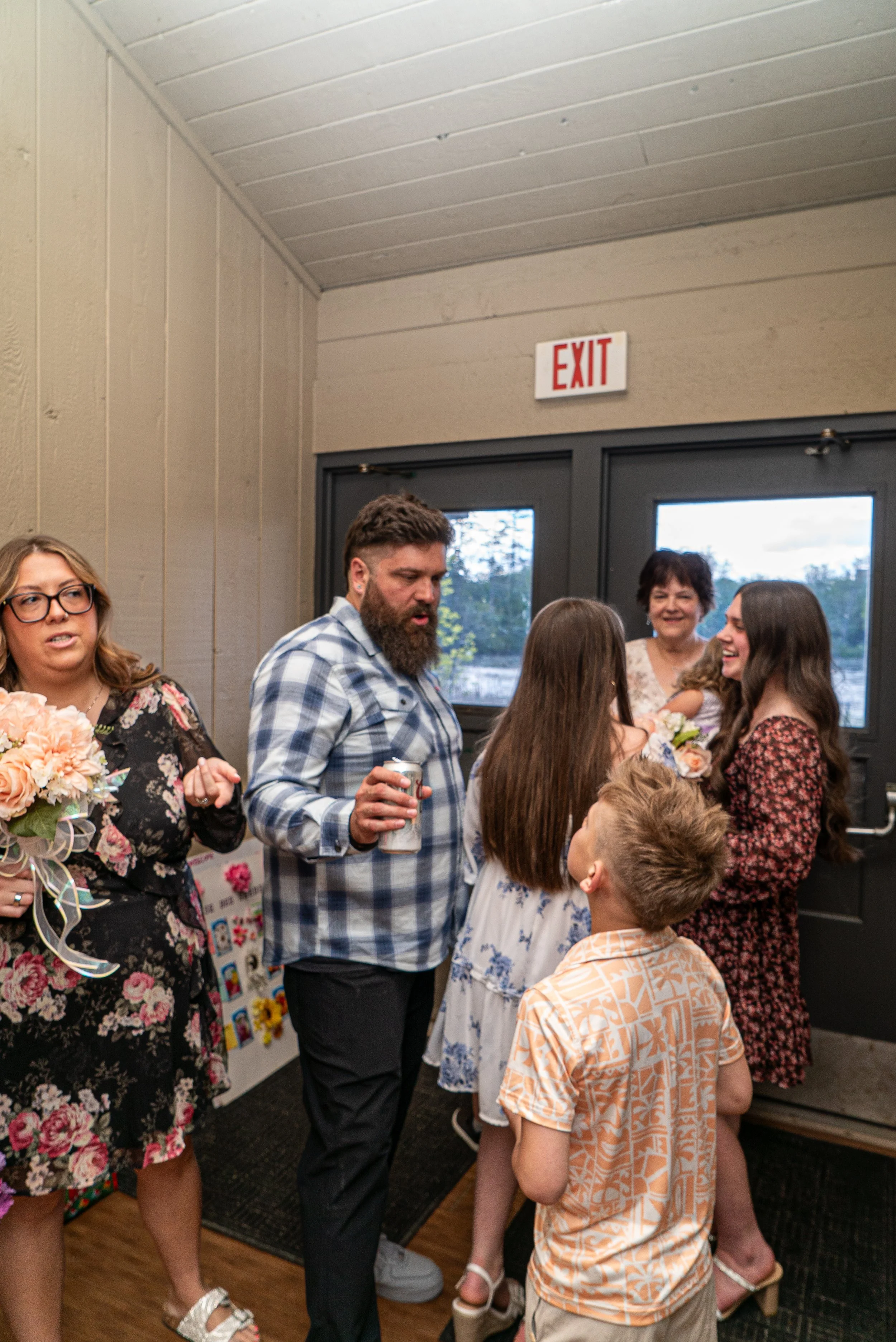 A group of people gathered indoors around a corner, engaged in conversation at a social event. There is a woman holding a bouquet of flowers, a man with a beard holding a drink, and children and adults talking and smiling.