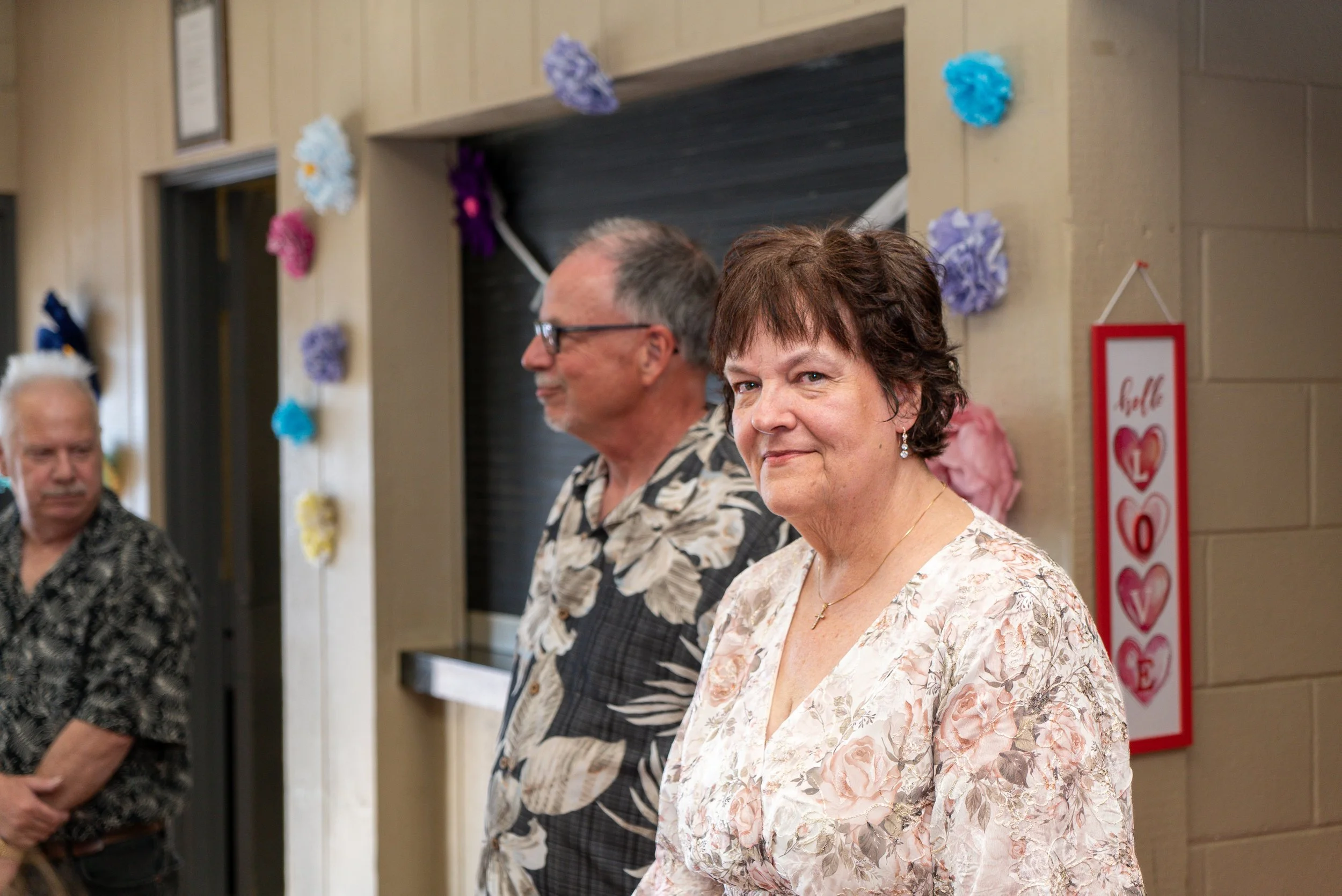 A group of elderly people standing in a row at a social event, with a woman in the foreground smiling, decorated with colorful paper flowers and a Valentine’s Day themed sign in the background.