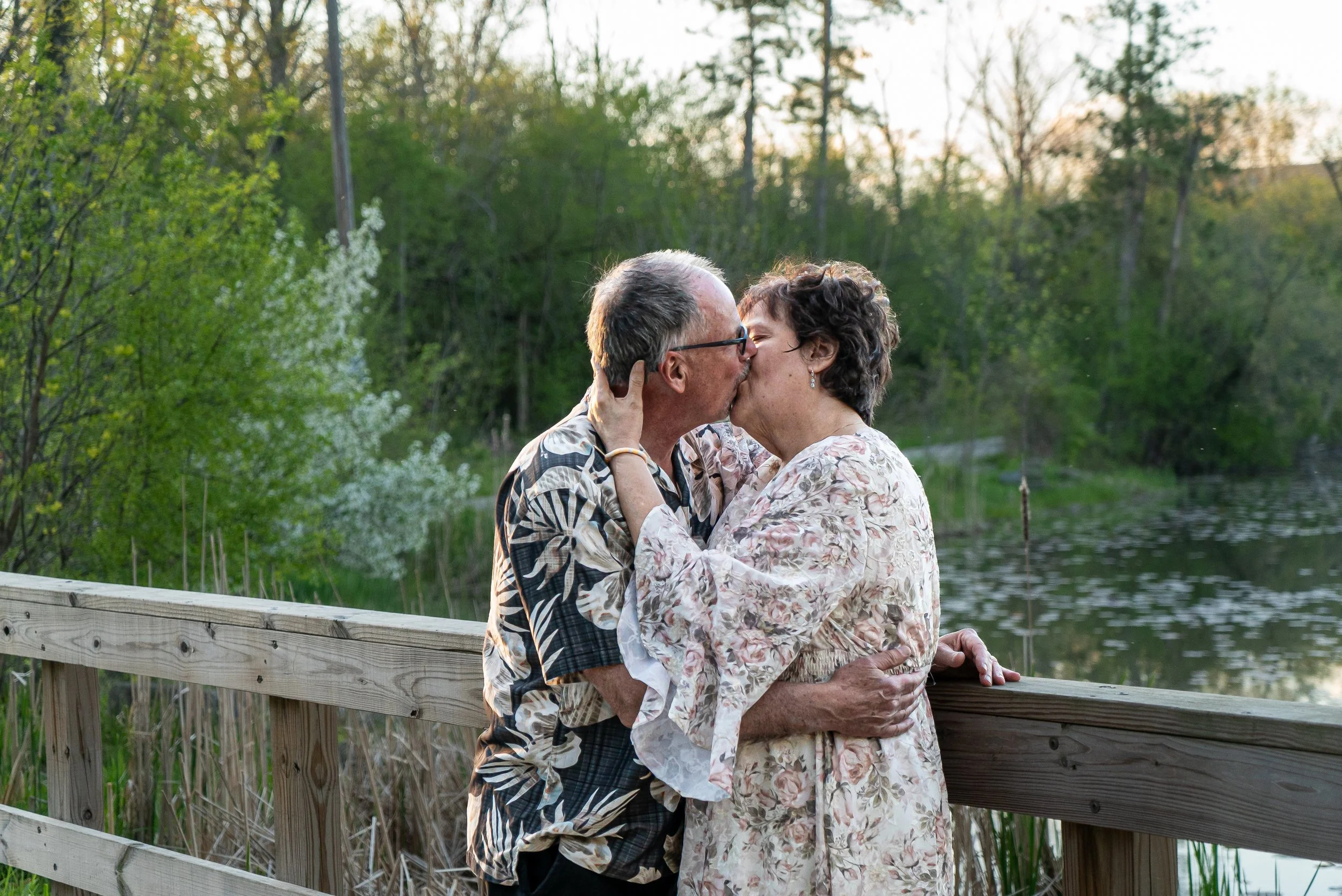 An elderly couple sharing a kiss on a wooden bridge by a pond, surrounded by green trees and foliage in the evening.