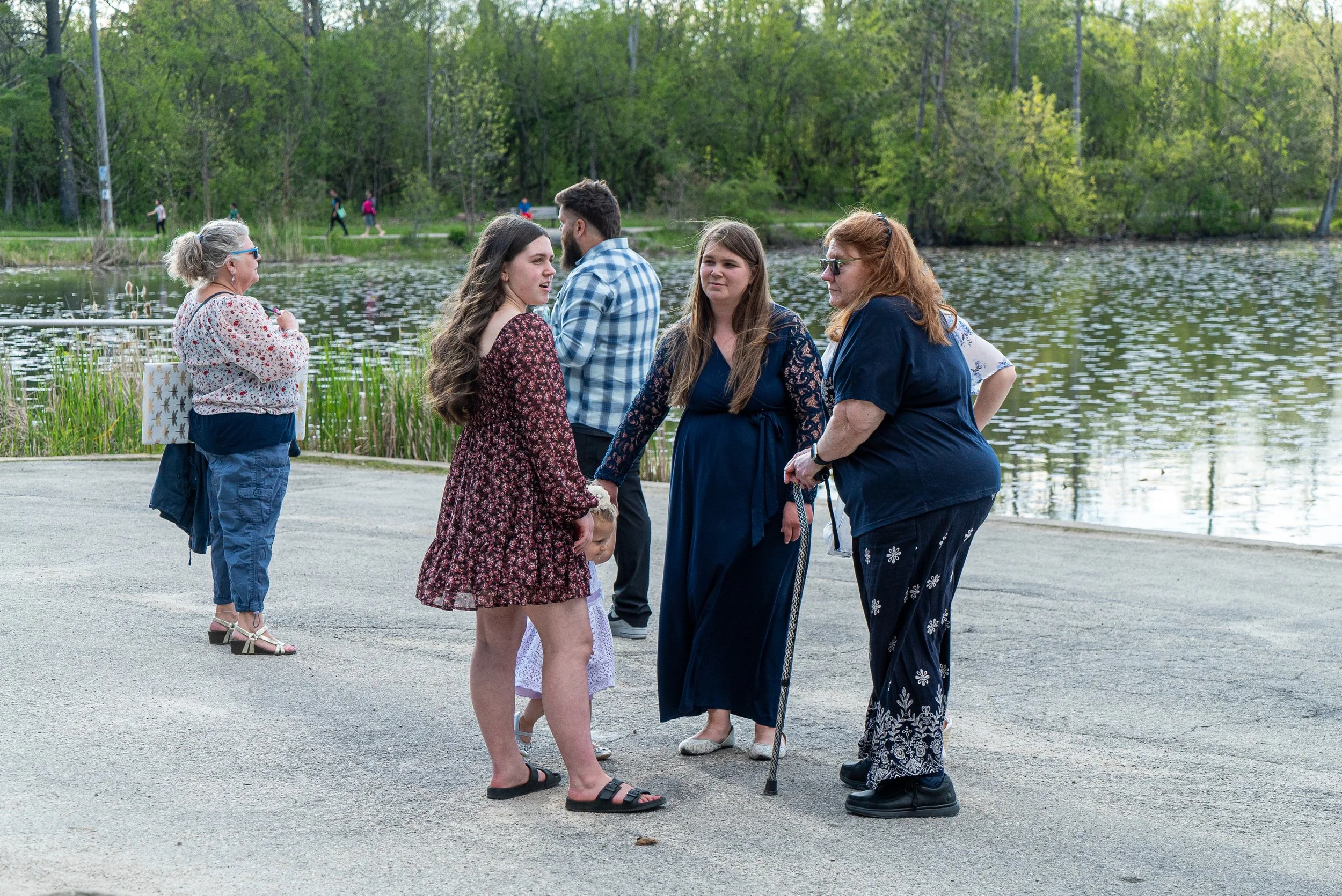A group of people by a lake, with a young woman and an older woman holding hands in the foreground, and others standing nearby, surrounded by trees and a pathway with people walking in the background.