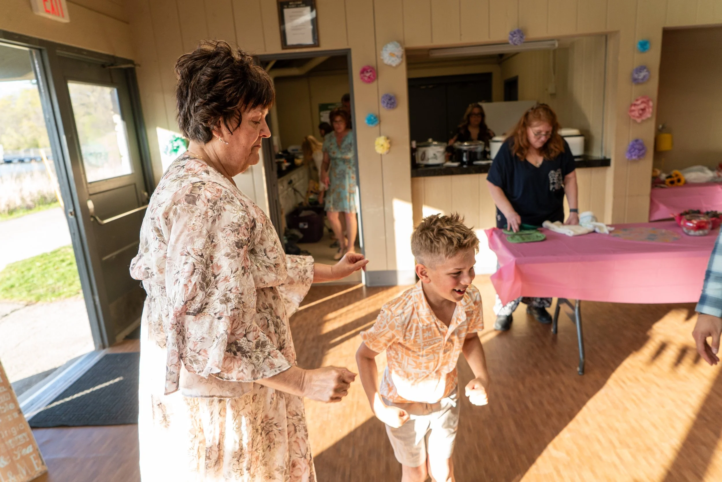 A group of women and children are gathered indoors at a party or celebration, with some standing and others sitting around a table covered with pink tablecloth, decorated with flowers and treats. One woman in a floral dress and a young boy in a light