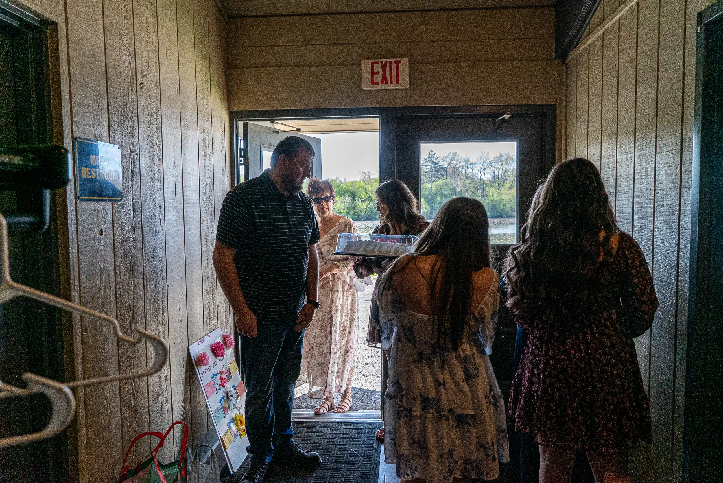 People standing at the entrance of a building near a window, with an open door leading outside where trees can be seen. A sign on the wall indicates the restroom and there is an EXIT sign above the door.